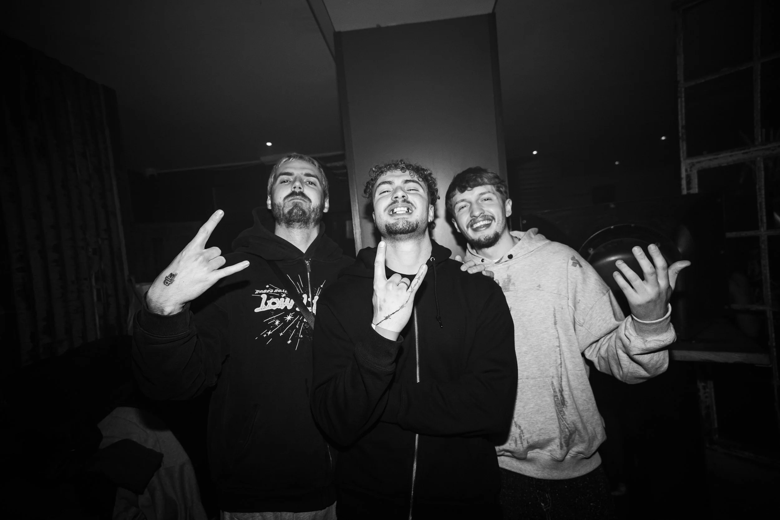 Trois jeunes hommes souriants posent ensemble, faisant des gestes de la main, dans un environnement intérieur sombre.
