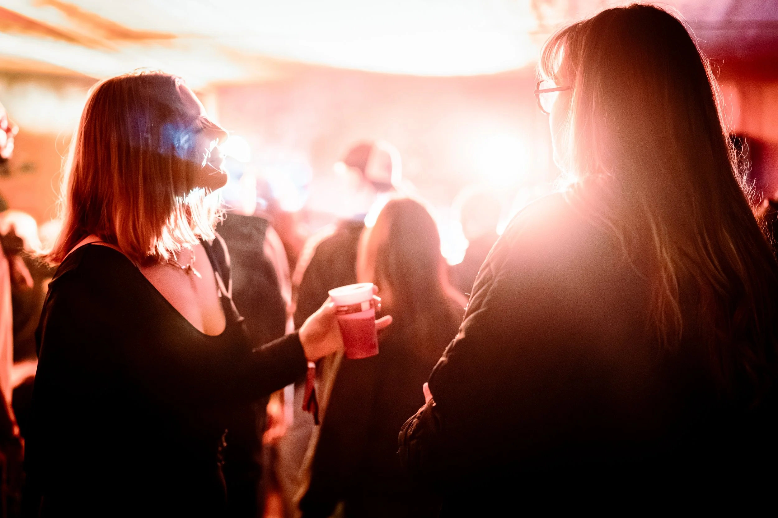 Deux jeunes femmes discutent dans une fête ou un événement, une d'elles tient un verre rouge, ambiance chaleureuse et lumineuse avec des personnes en arrière-plan.
