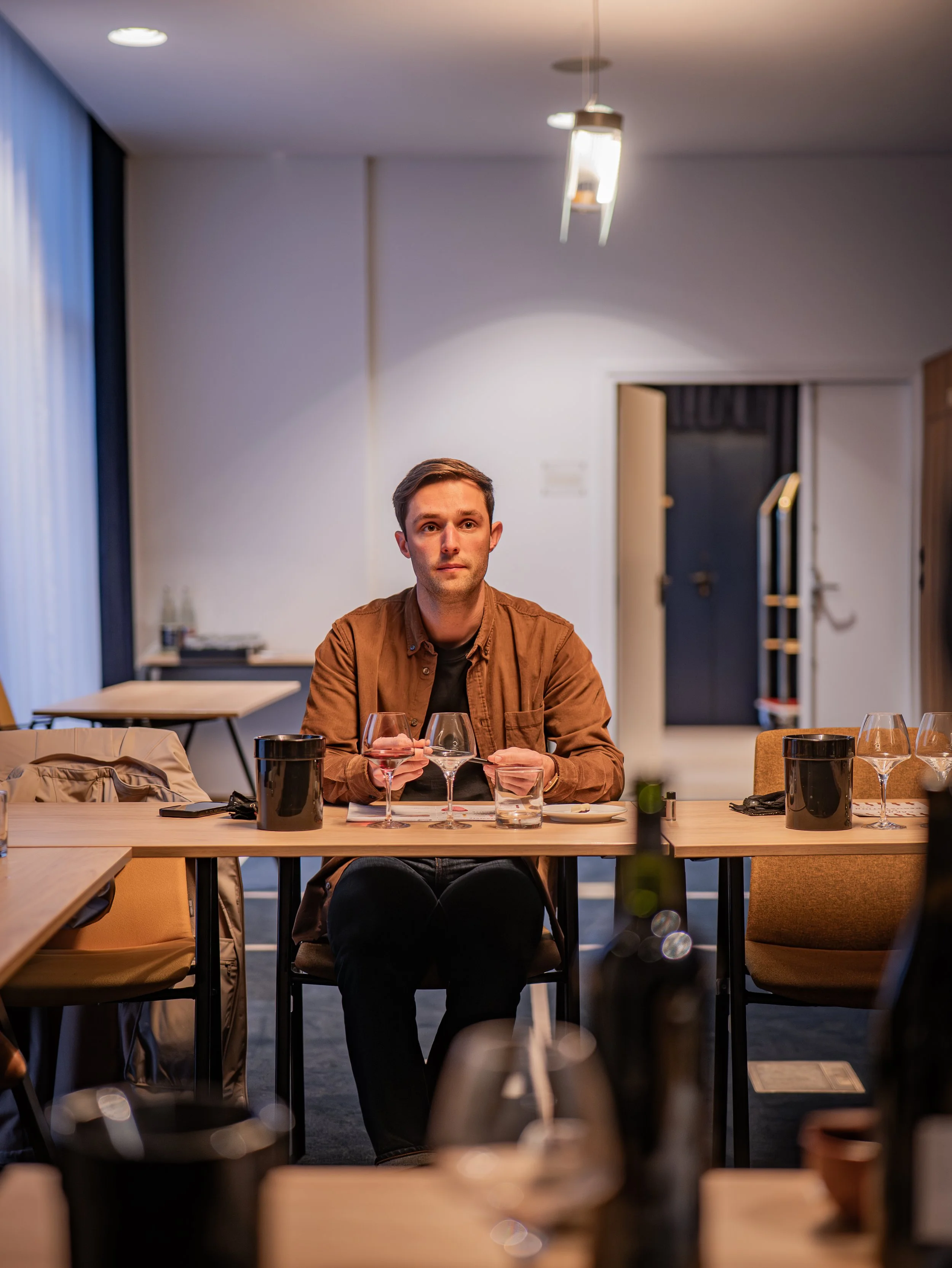 Un homme assis à une table dans un restaurant, tenant un verre de vin rouge, avec d'autres verres et bouteilles sur la table, dans un environnement moderne avec éclairage tamisé.