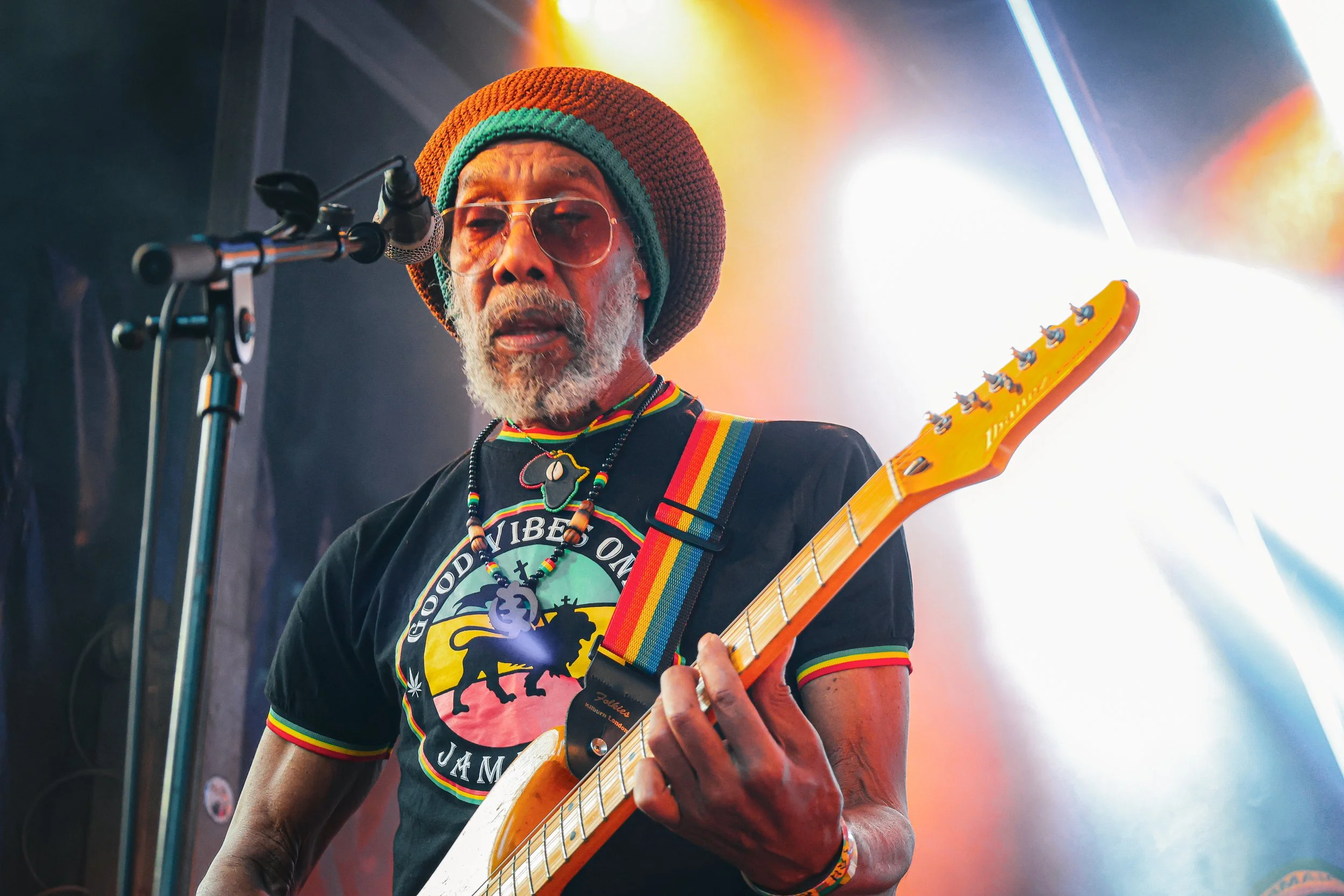 Un homme jouant de la guitare en concert, portant un chapeau coloré et des lunettes, avec un fond lumineux coloré.