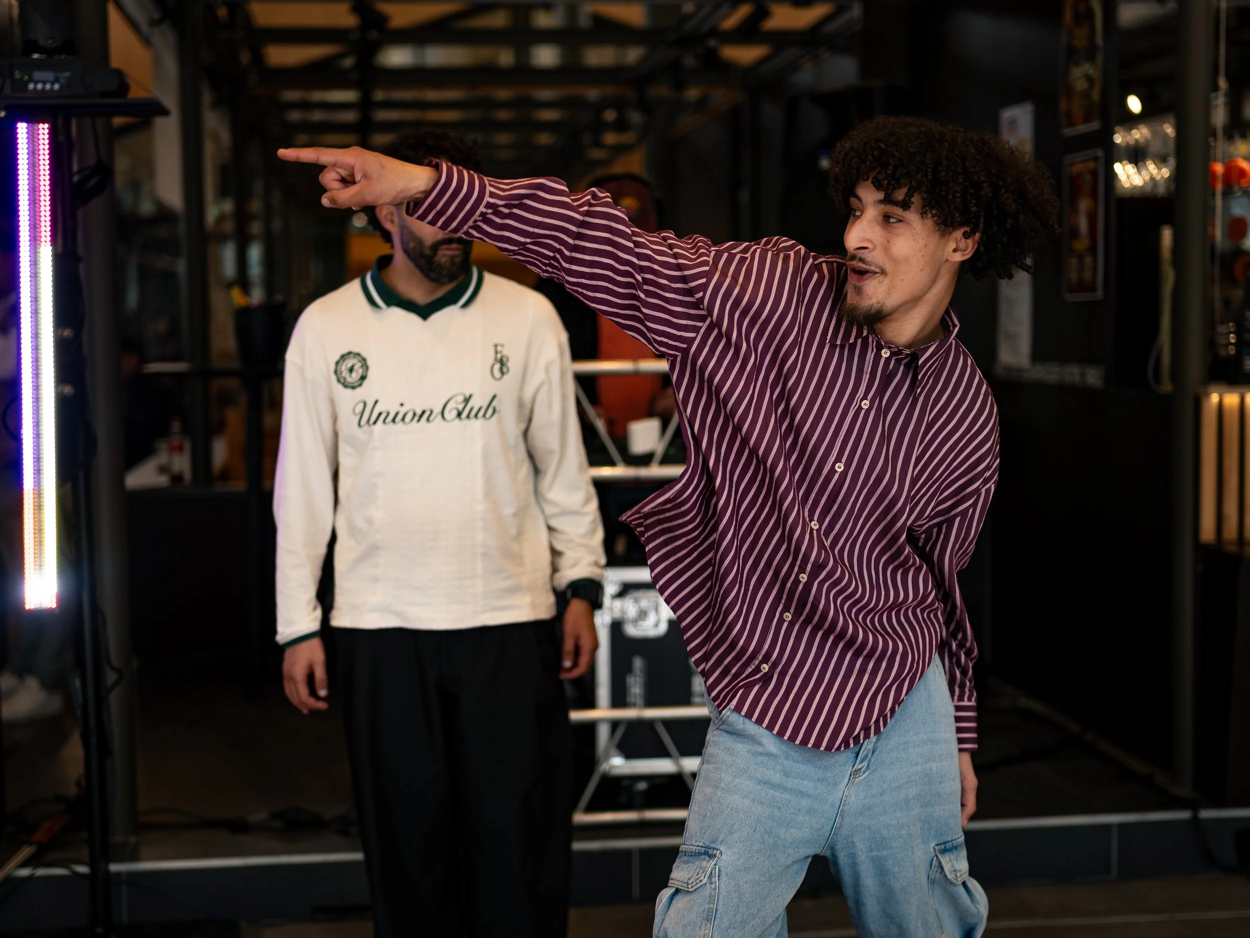 Homme jeune avec des cheveux bouclés portant une chemise à rayures violettes et des jeans bleus, faisant un geste de pointage, dans un endroit intérieur avec des personnes en arrière-plan.