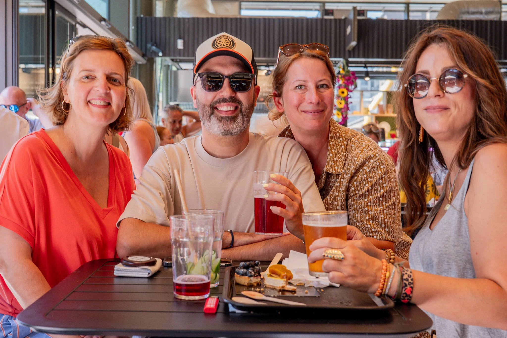Groupe de quatre personnes assises à une table au restaurant, mangeant et buvant, souriant et portant des lunettes de soleil, avec des boissons et de la nourriture sur la table.