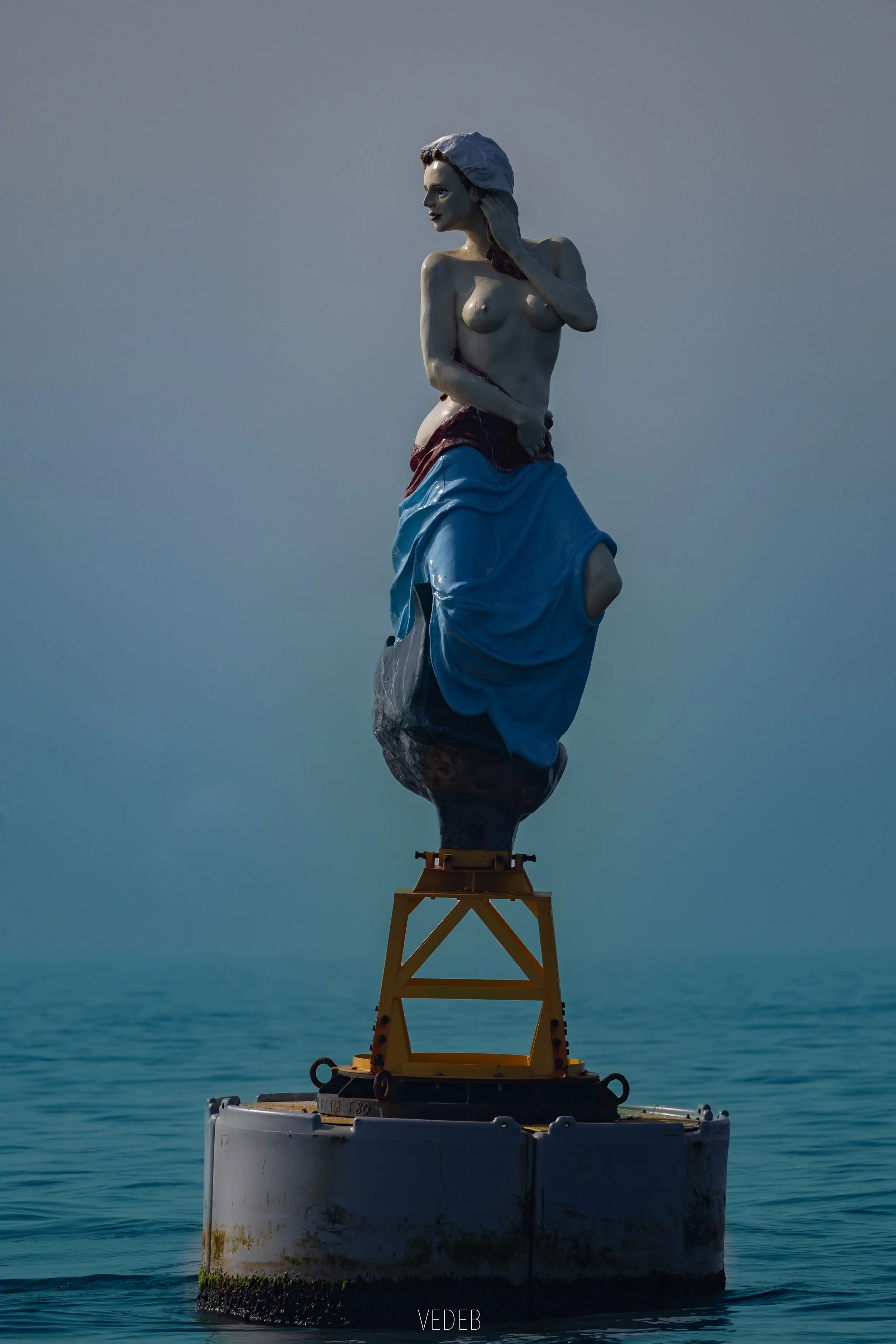 Une statue en forme de femme nue tenant un tissu, placée sur une plateforme flottante dans la mer, contre un ciel dégagé.
