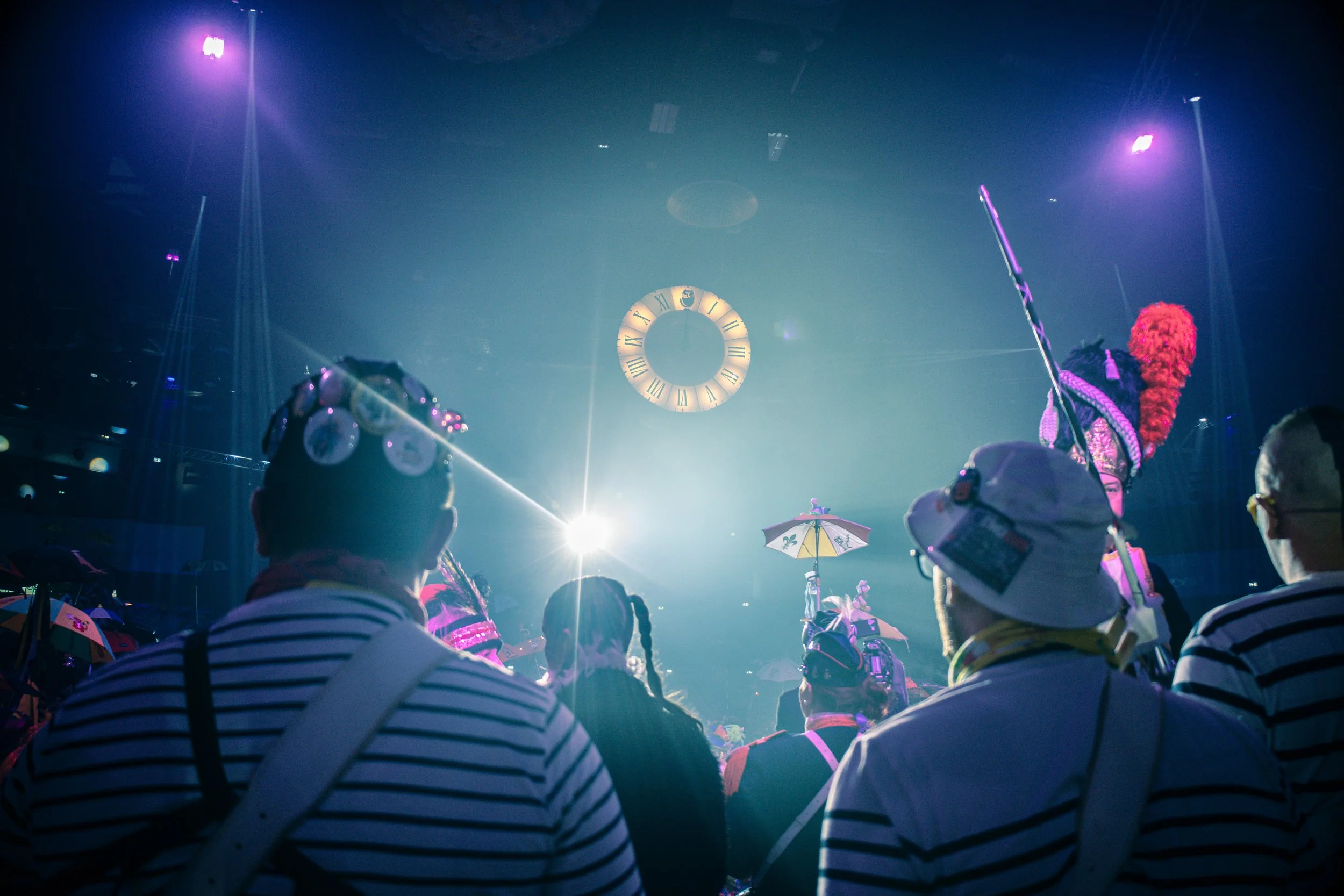 Une foule de personnes portant des vêtements rayés, un cercle lumineux à l'arrière-plan, un grand horloge circulaire avec des chiffres romains suspendu au plafond, plusieurs projecteurs violets et des parapluies colorés dans une ambiance de spectacle
