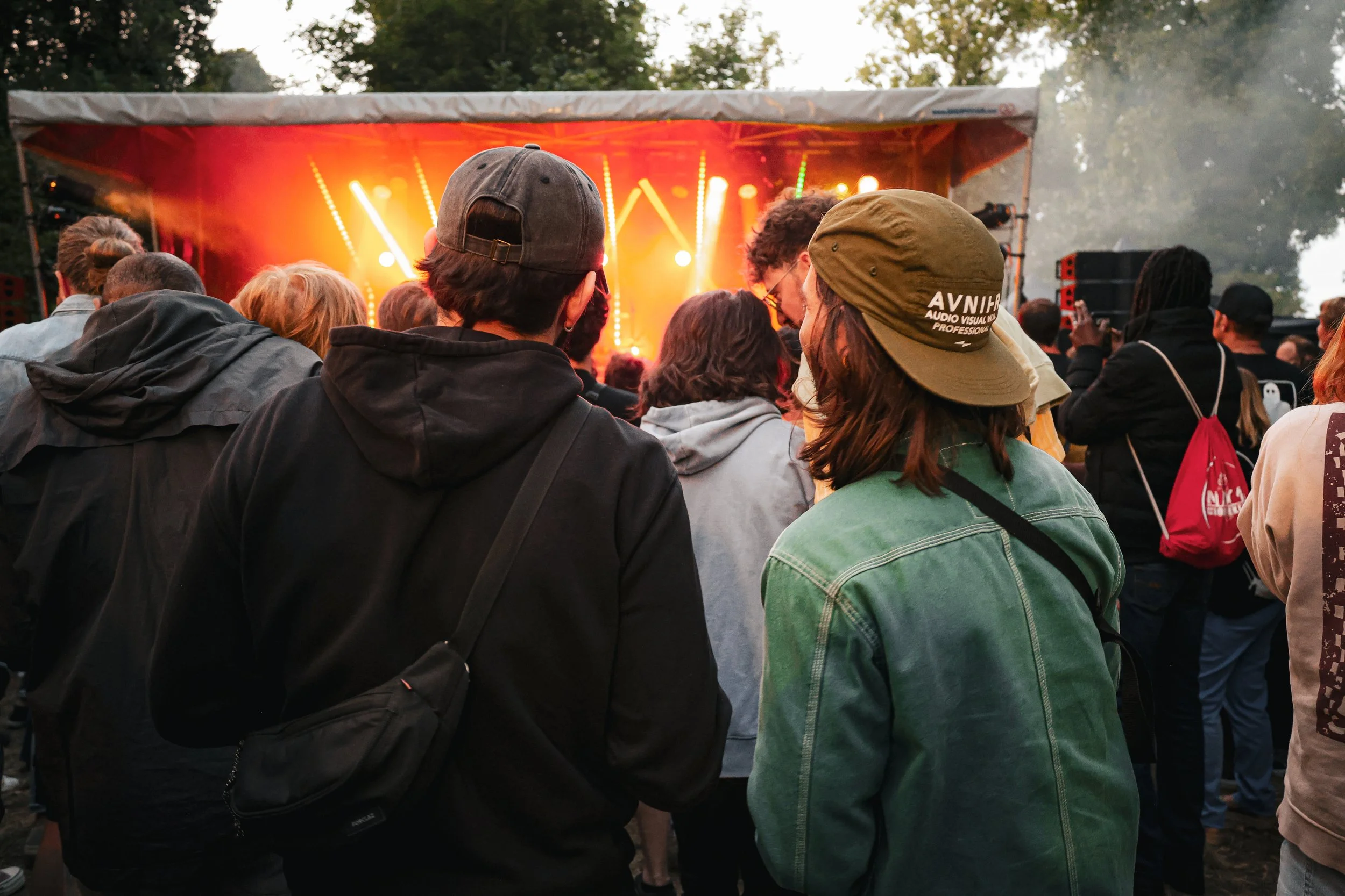 Un groupe de personnes regardant un concert en plein air avec des lumières colorées sur scène. Certaines personnes portent des casquettes et des sacs à dos.