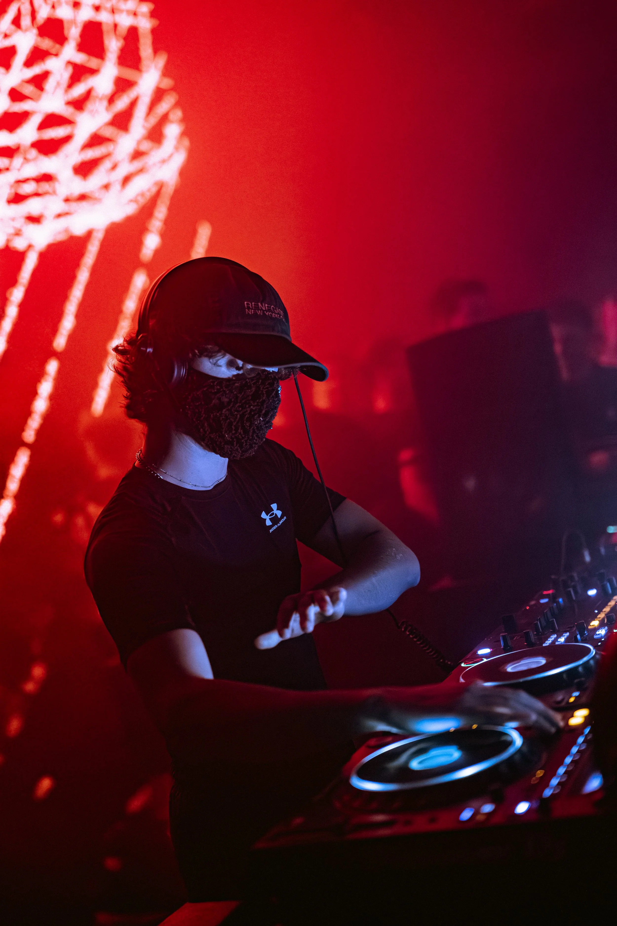 DJ portant un masque, un casque et un T-shirt noir, mixant à une soirée dans une ambiance rouge avec des lumières et un écran lumineux en arrière-plan.