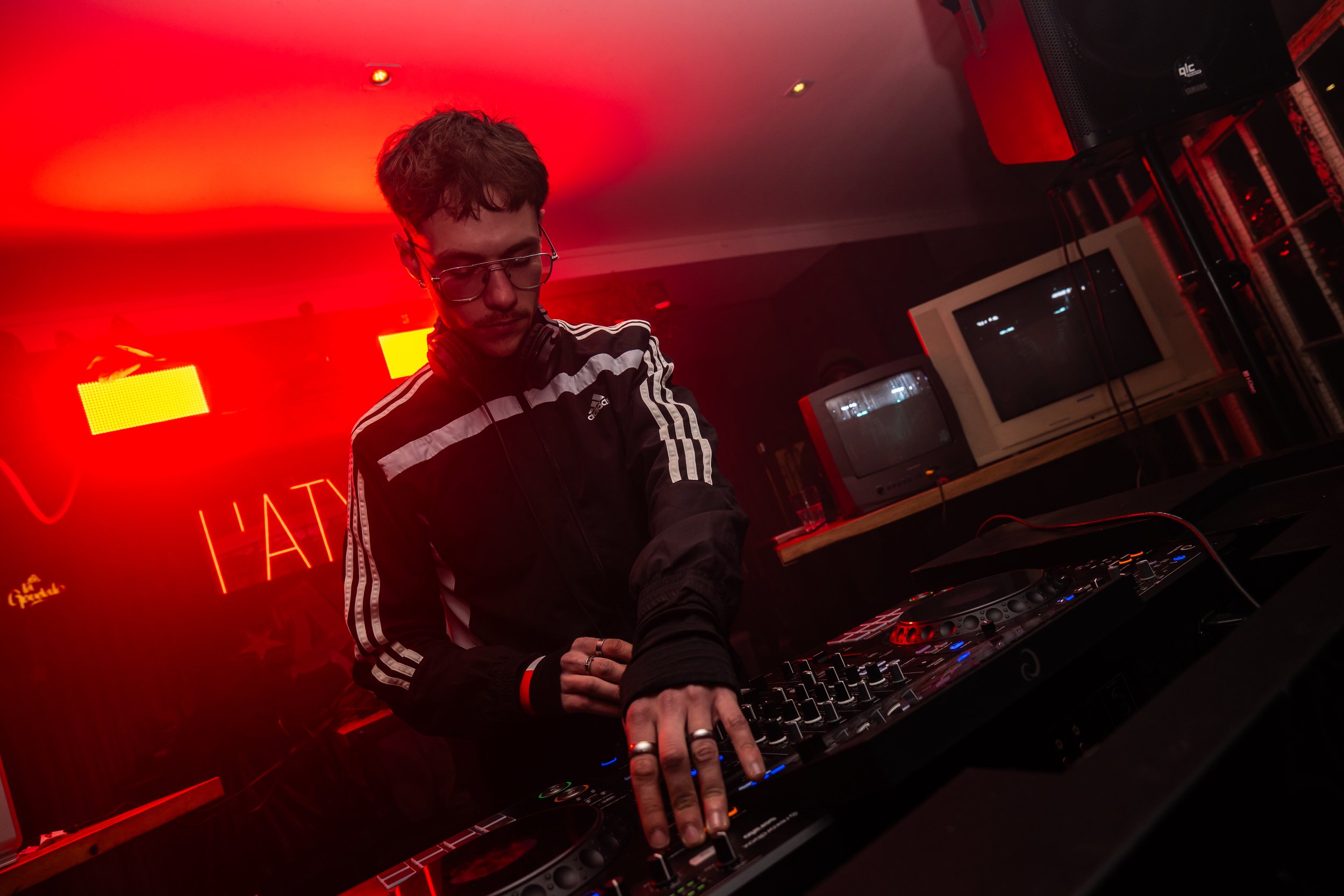 Un DJ portant des lunettes de soleil joue derrière un DJ set, entouré d'une ambiance de soirée avec un éclairage rouge et des écrans lumineux.