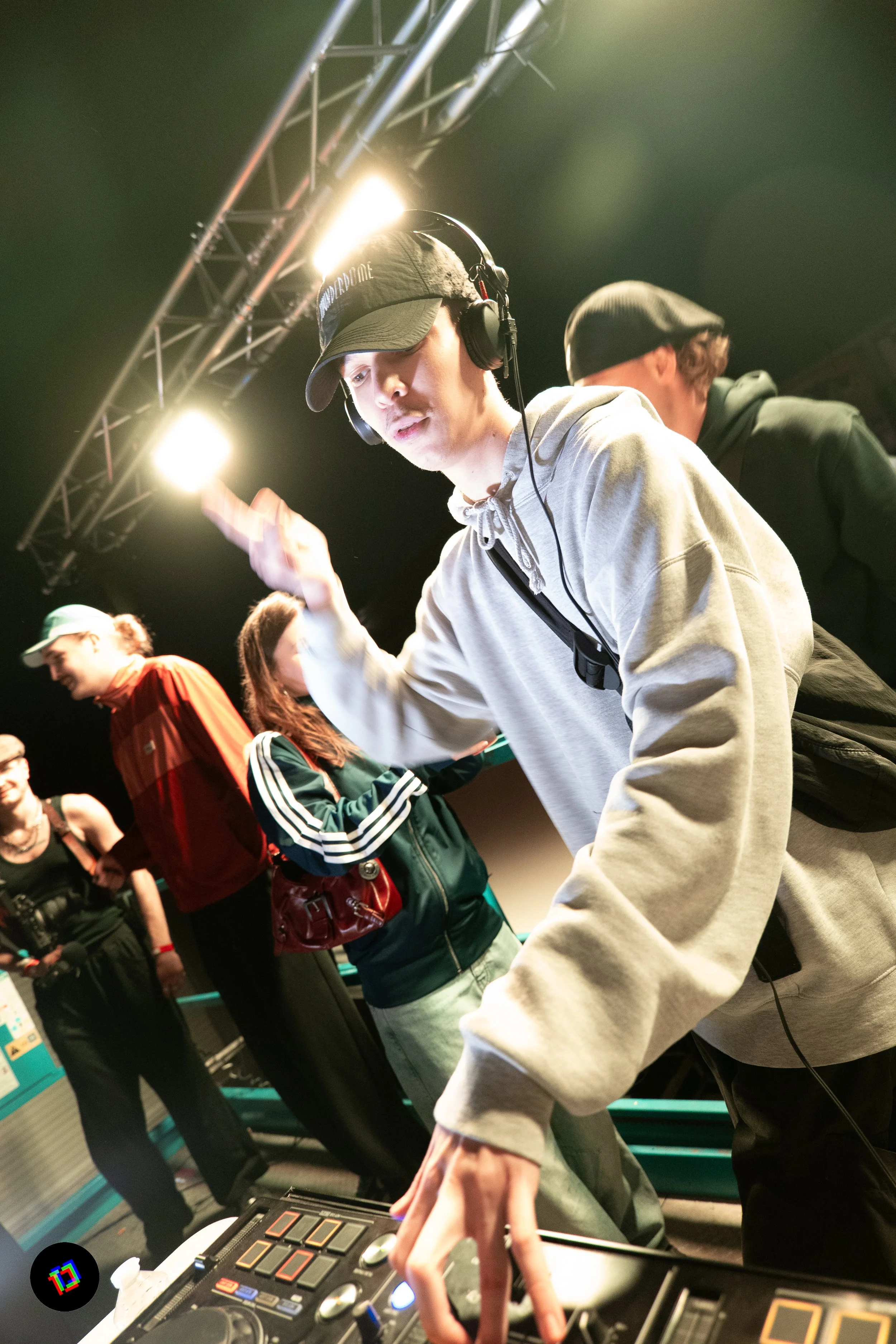 Un jeune DJ portant un casque et un bonnet, manipulant des équipements de sonorisation lors d'un événement. Il y a plusieurs personnes en arrière-plan, dans une ambiance sombre avec des lumières vives.