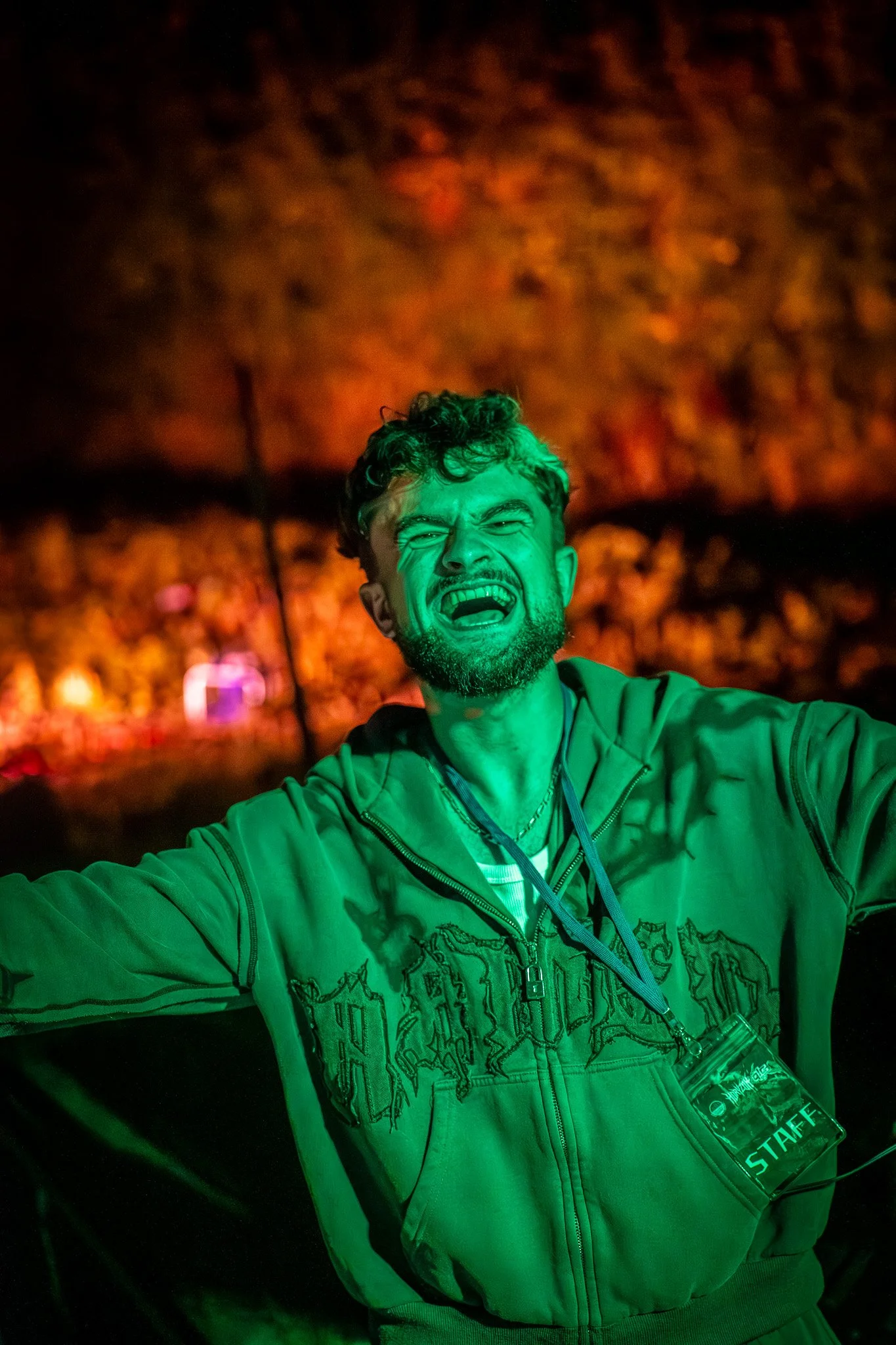 Jeune homme avec un sourire éclatant, portant une veste verte, dans un environnement avec un éclairage coloré, probablement lors d'une soirée ou d'un événement, avec un badge d'identification autour du cou indiquant qu'il est un membre du personnel.