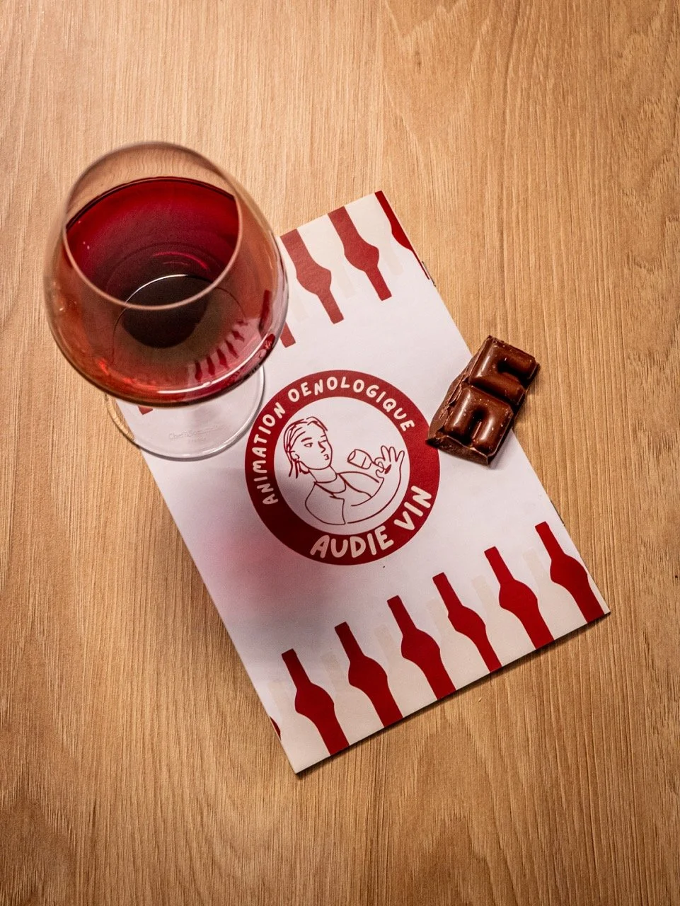Verre de vin rosé, tablette de chocolat et une affiche avec le logo d'AUDIF, une école d'animation et d'œnologie, sur une table en bois.