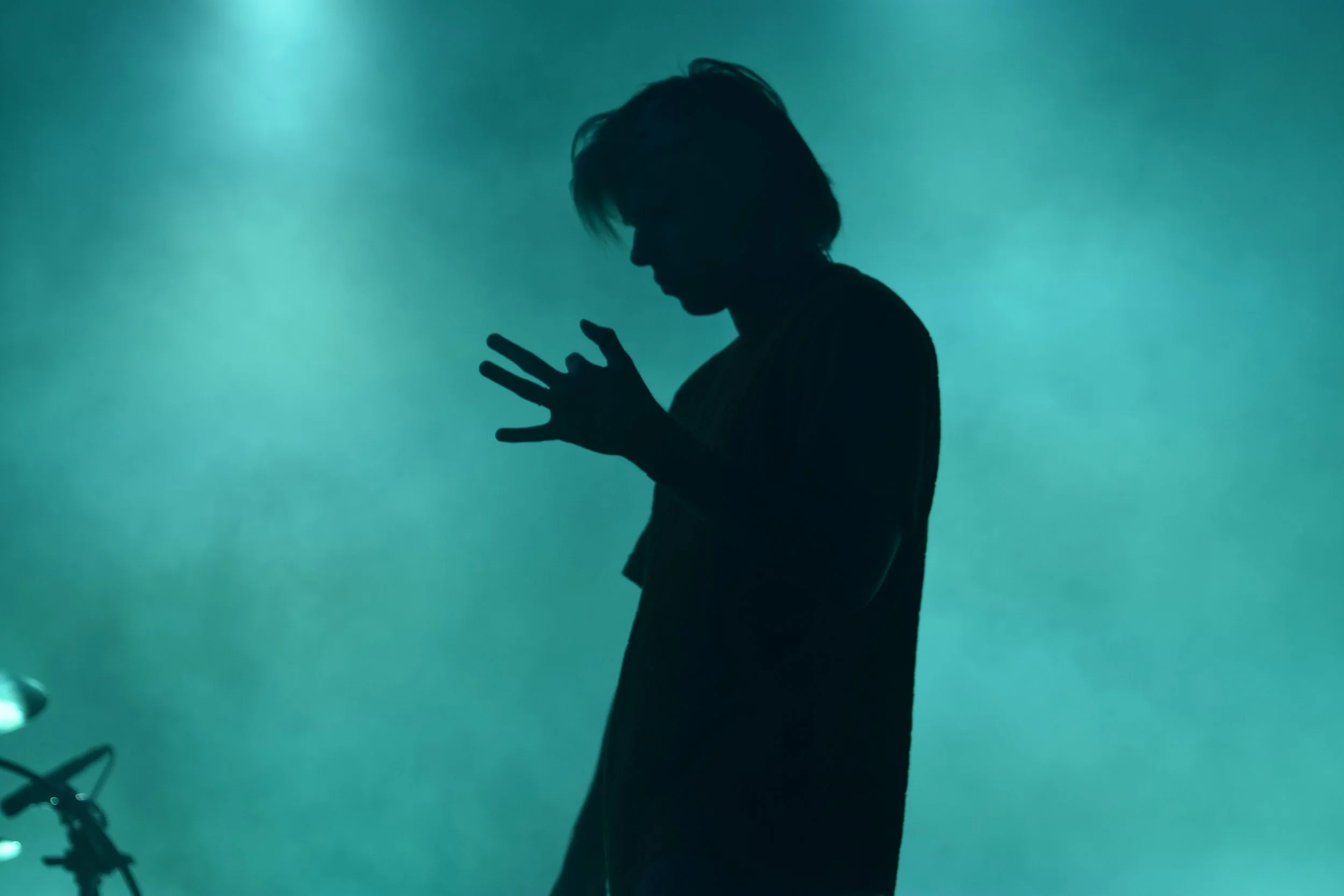 Silhouette d'une personne, probablement un homme, avec des cheveux longs, debout dans une pièce sombre avec un fond vert. La personne tient peut-être un objet ou une main sur sa poitrine.