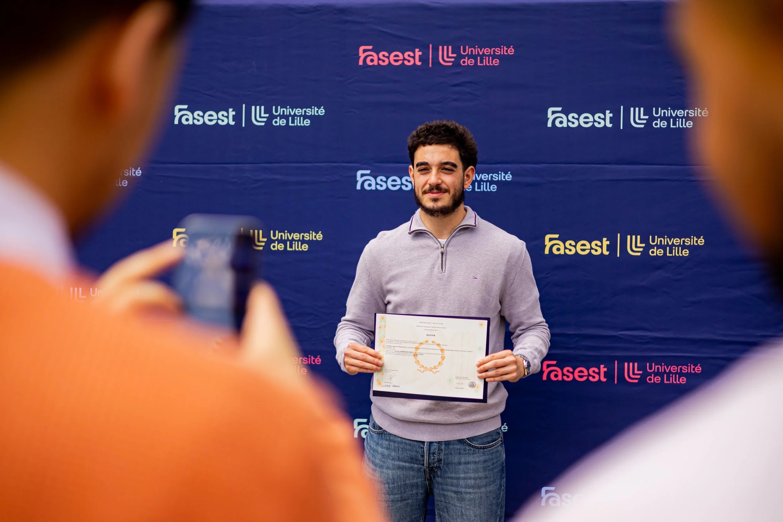 Une jeune homme tient un certificat lors d'une cérémonie de remise de diplôme devant un fond avec le logo de l'Université de Lille et FASET.