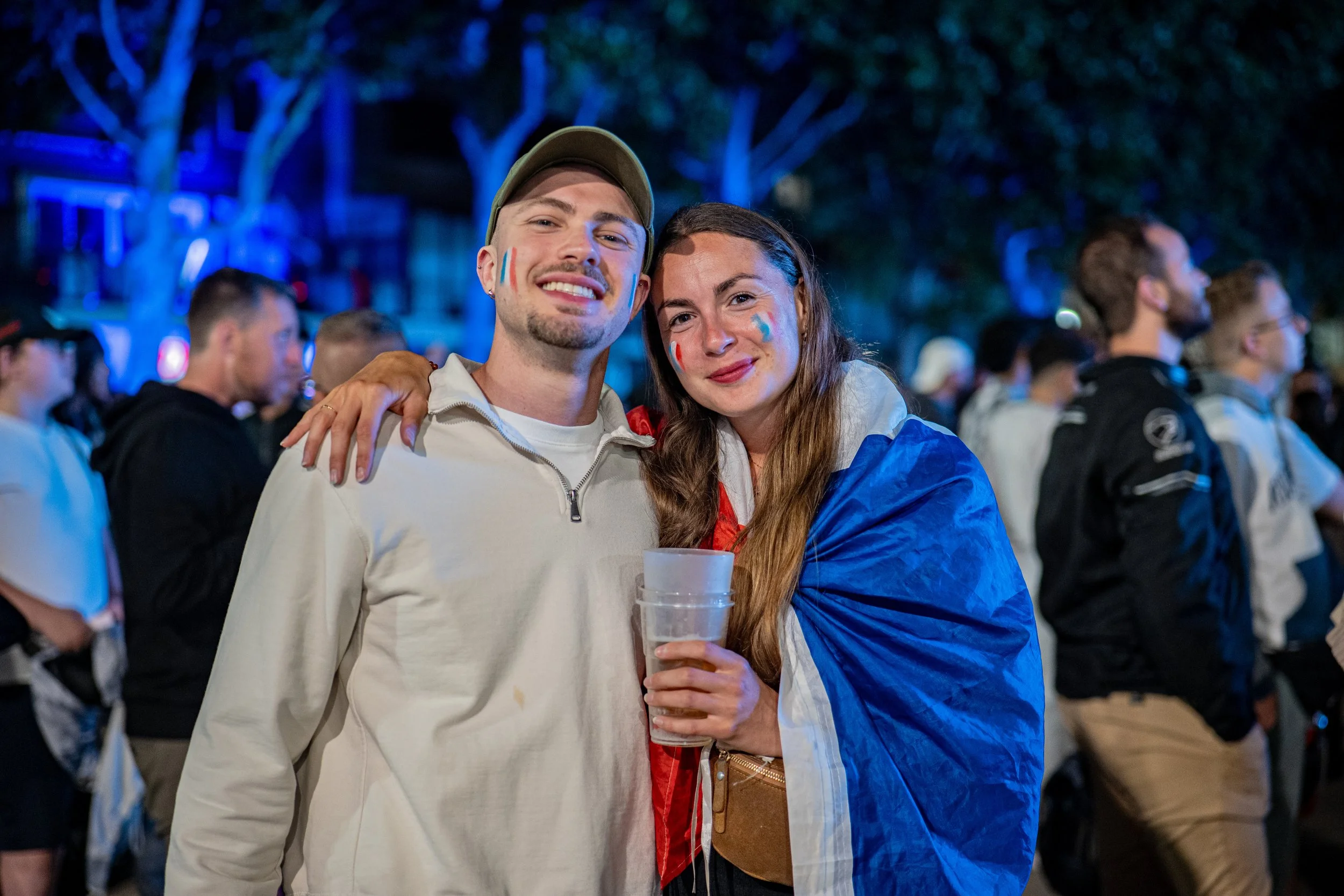 Deux personnes souriantes se tiennent près l'une de l'autre lors d'un événement en soirée, l'une d'elles porte un drapeau français peint sur la joue. Ils tiennent un verre de bière, et il y a un groupe de spectateurs en arrière-plan. L'ambiance est f