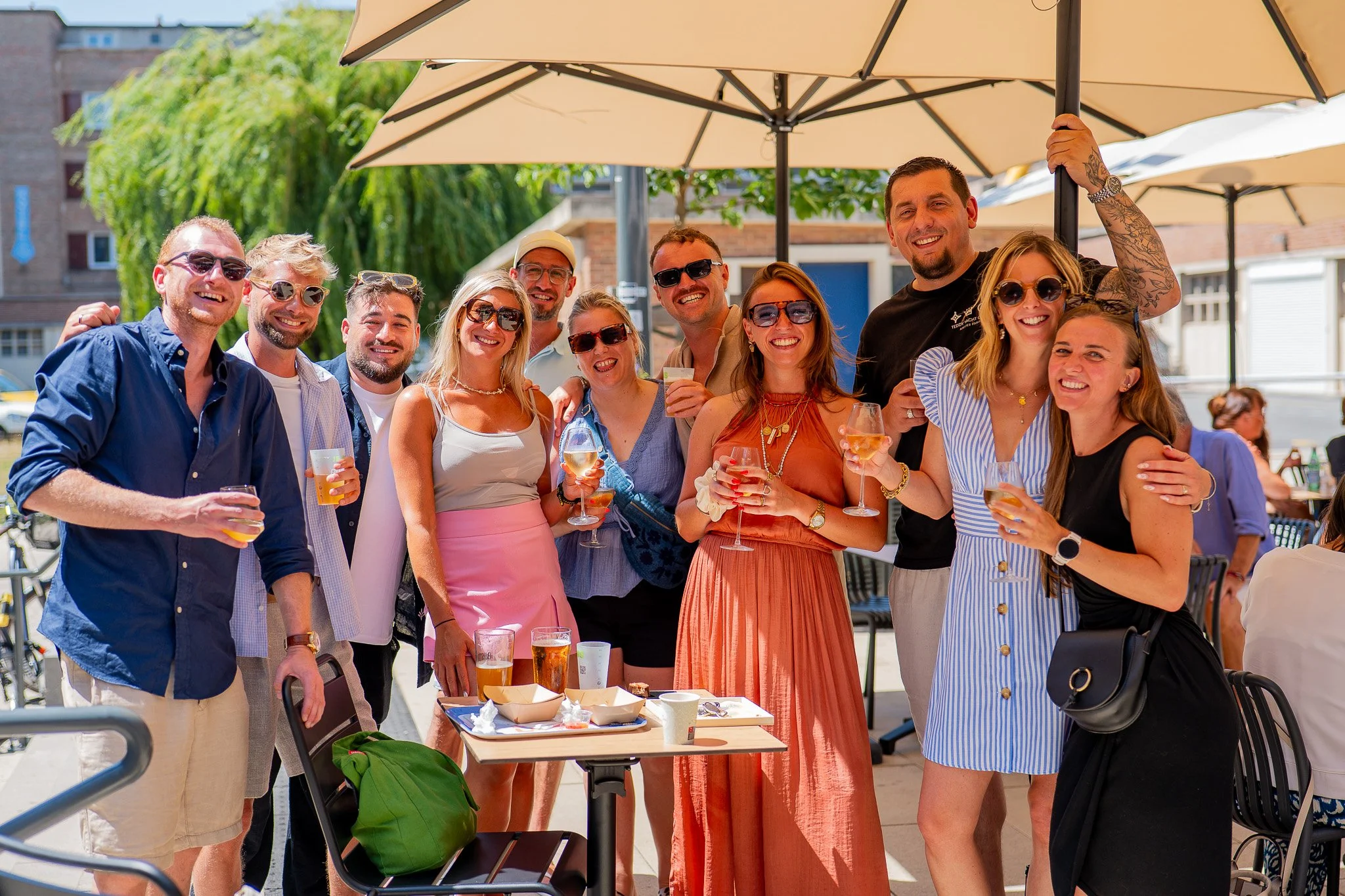Groupe de personnes profitant d'un rassemblement en plein air avec parasols, tenant des verres de boisson, souriant et posant pour la photo. Il y a une table avec des snacks et des boissons, ambiance ensoleillée.