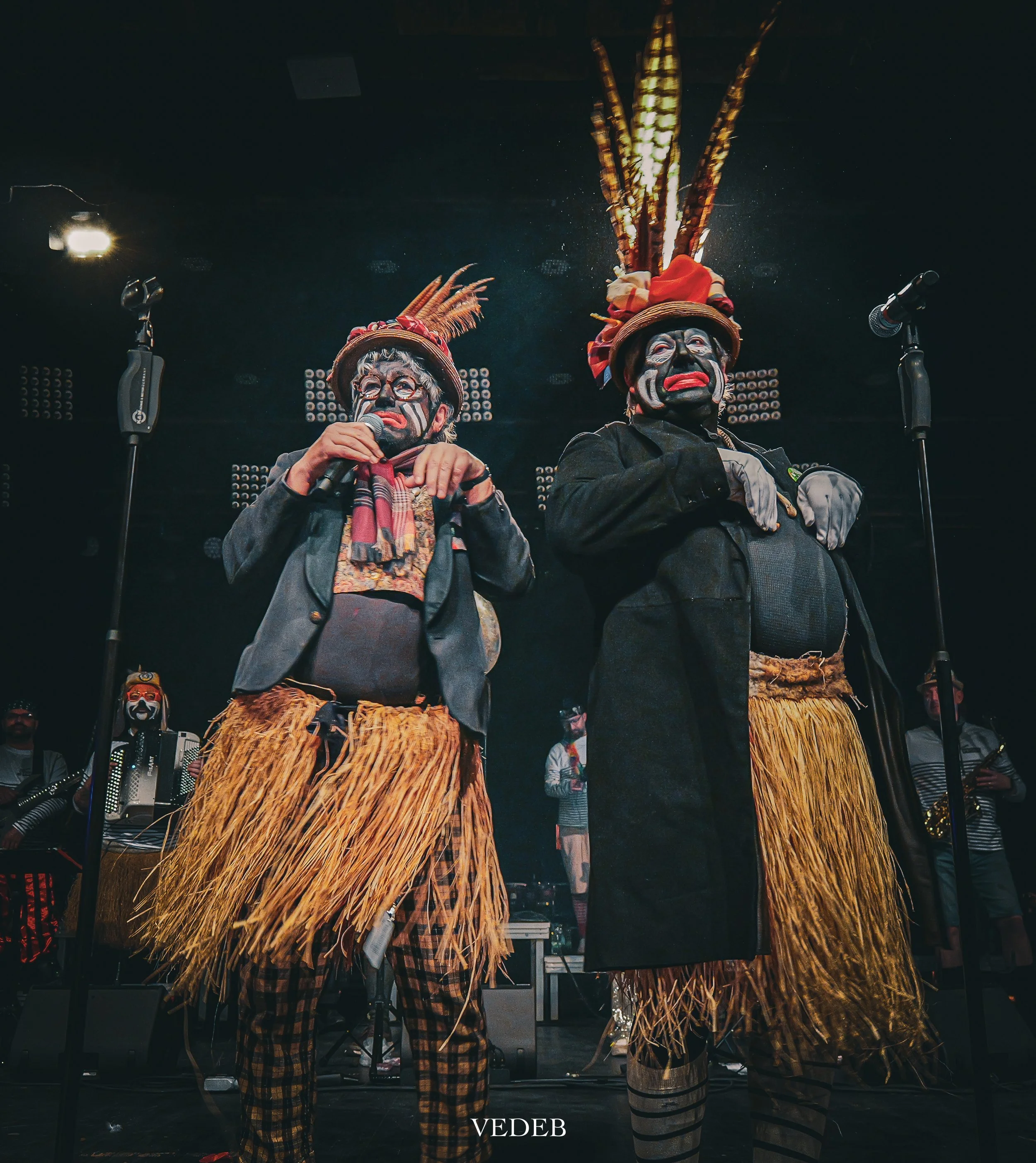 Deux personnes en costume traditionnel, portant des masques et des chapeaux ornés de plumes, lors d'une performance ou célébration, avec microphones et éclairage de scène.