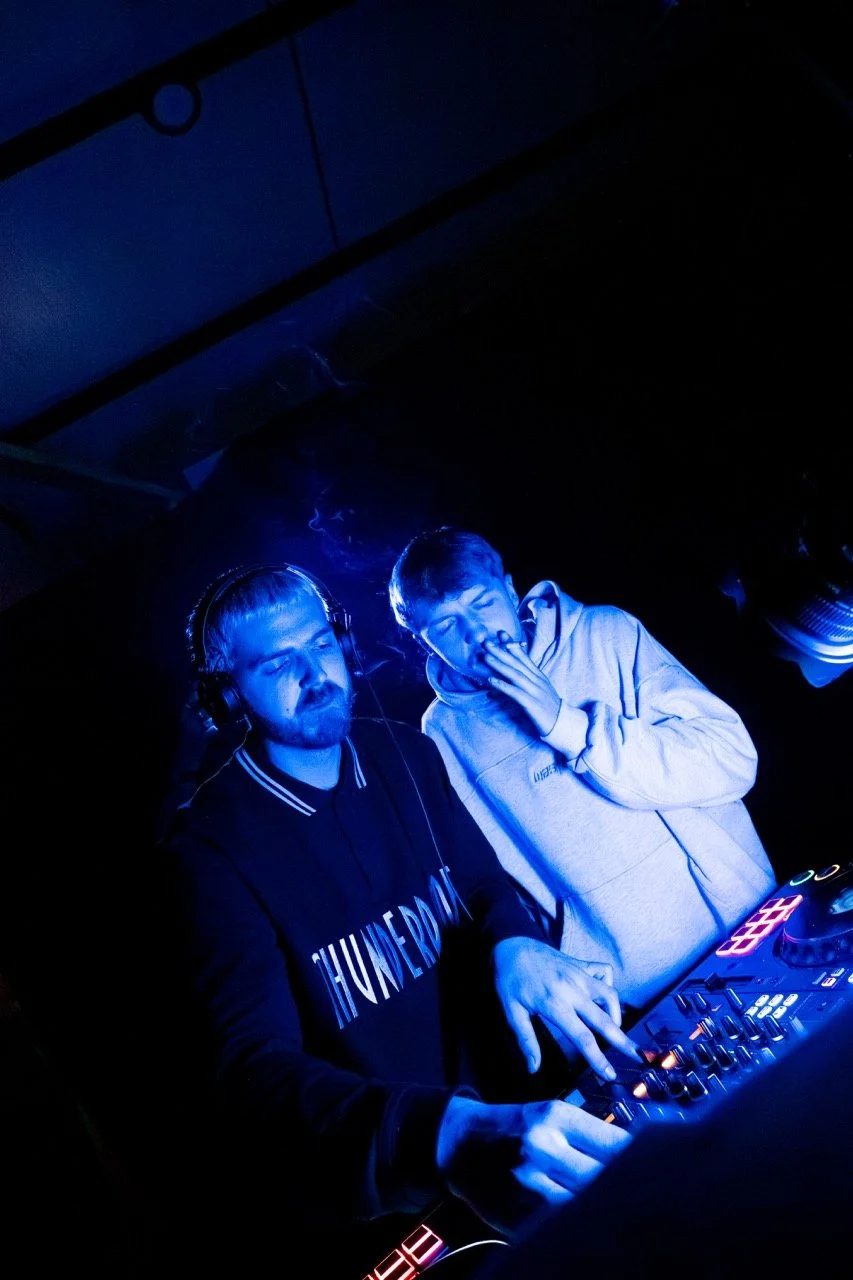 Deux jeunes hommes en train de mixer de la musique dans une boîte de nuit, éclairés par une lumière bleue.