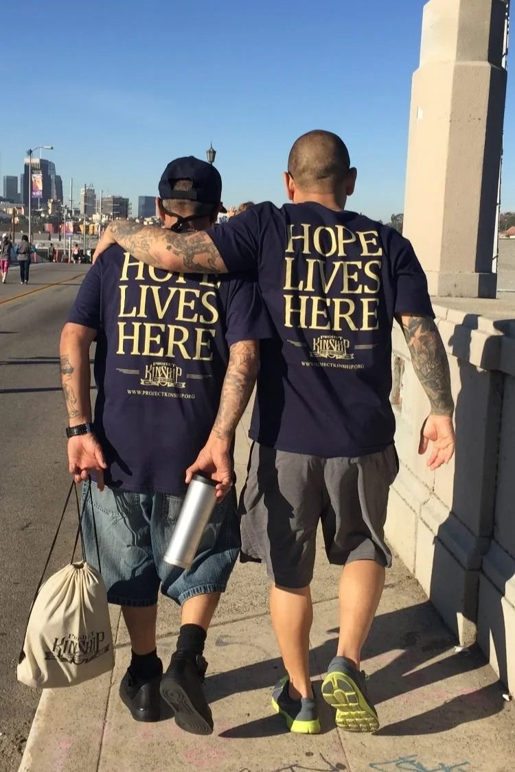 Two men with tattoos walking arm-in-arm on a city bridge, wearing matching navy blue t-shirts that say 'Hope LIVES HERE,' with one man carrying a beige drawstring bag and a silver water bottle.