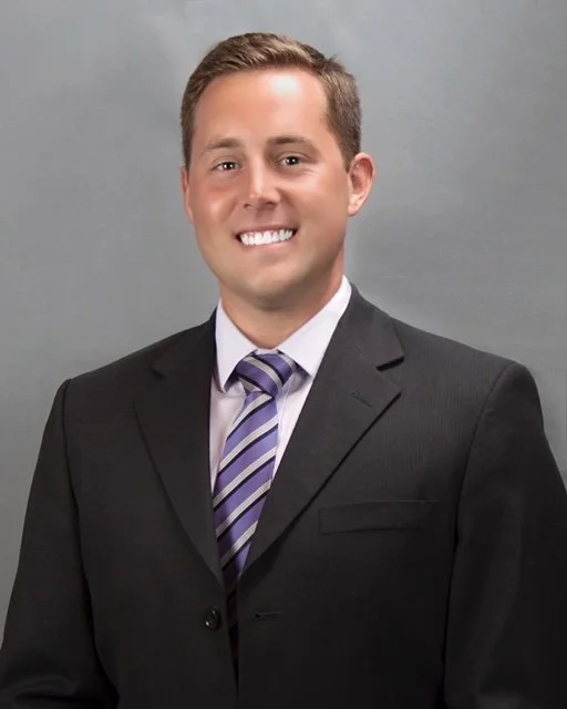 Professional headshot of a smiling man in a dark suit, white shirt, and striped tie, against a plain gray background.