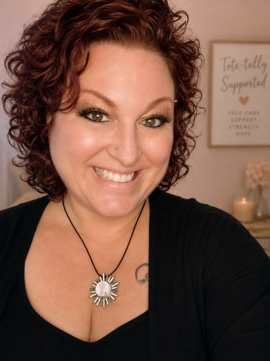 A smiling woman with short, curly red hair, wearing a black top and a necklace with a pink ribbon pendant. In the background, there is a sign that reads 'Totally supported' with words like self-care, support, strength, and hope, and some flowers and a candle.