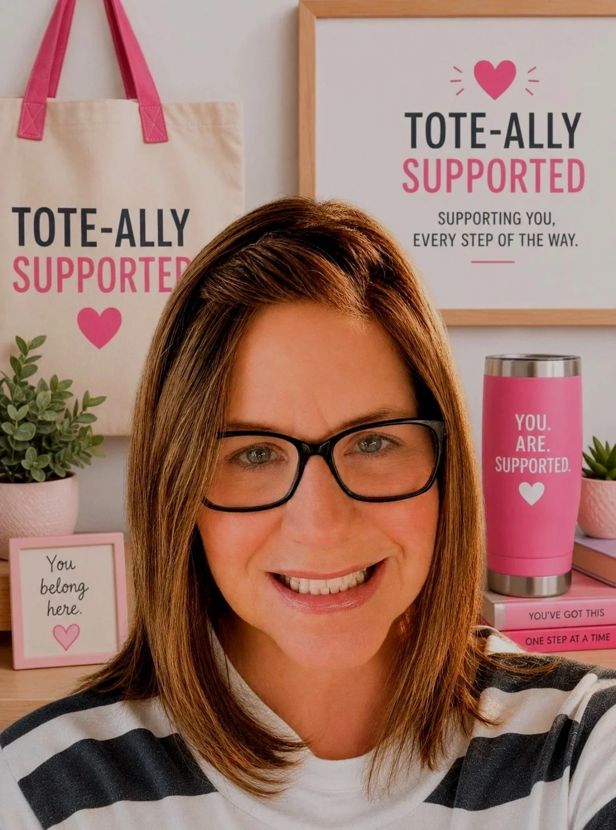 A woman with glasses smiling for a selfie in front of supportive tote bags, plants, and motivational signs featuring pink and white text.