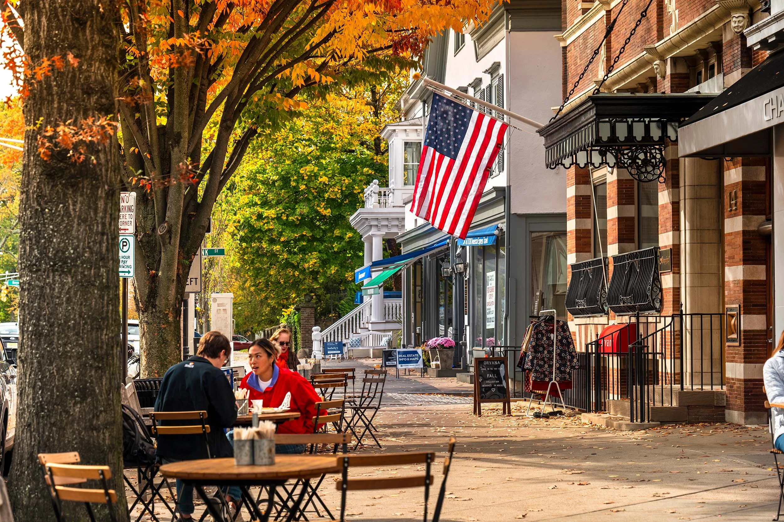 A lively street scene in autumn with trees showing orange and green leaves, cafes with outdoor seating and people dining, a red-brick building and an American flag hanging outside.