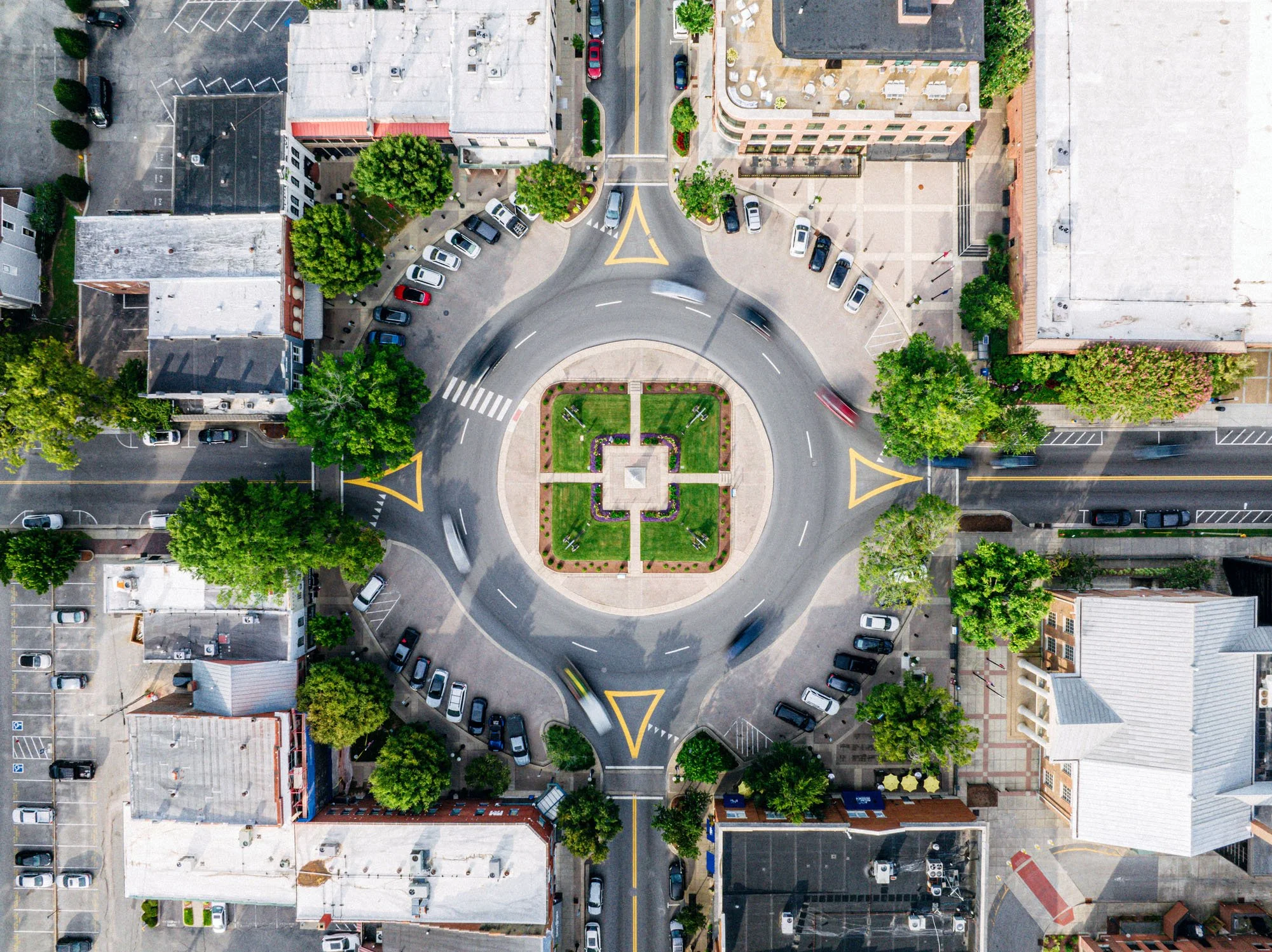 Aerial view of a city intersection with a roundabout, surrounding buildings, parking lots, and trees.