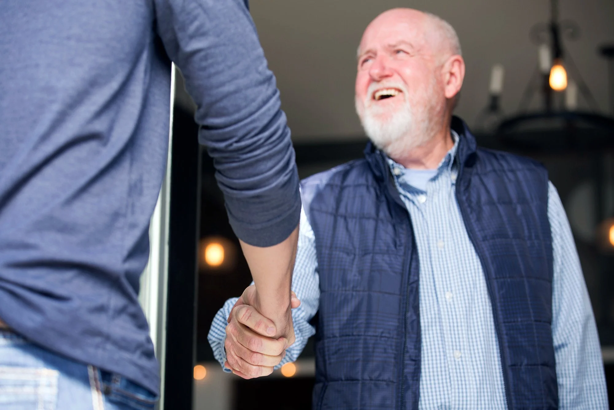An elderly man with a white beard smiling as he shakes hands and greets a younger person.