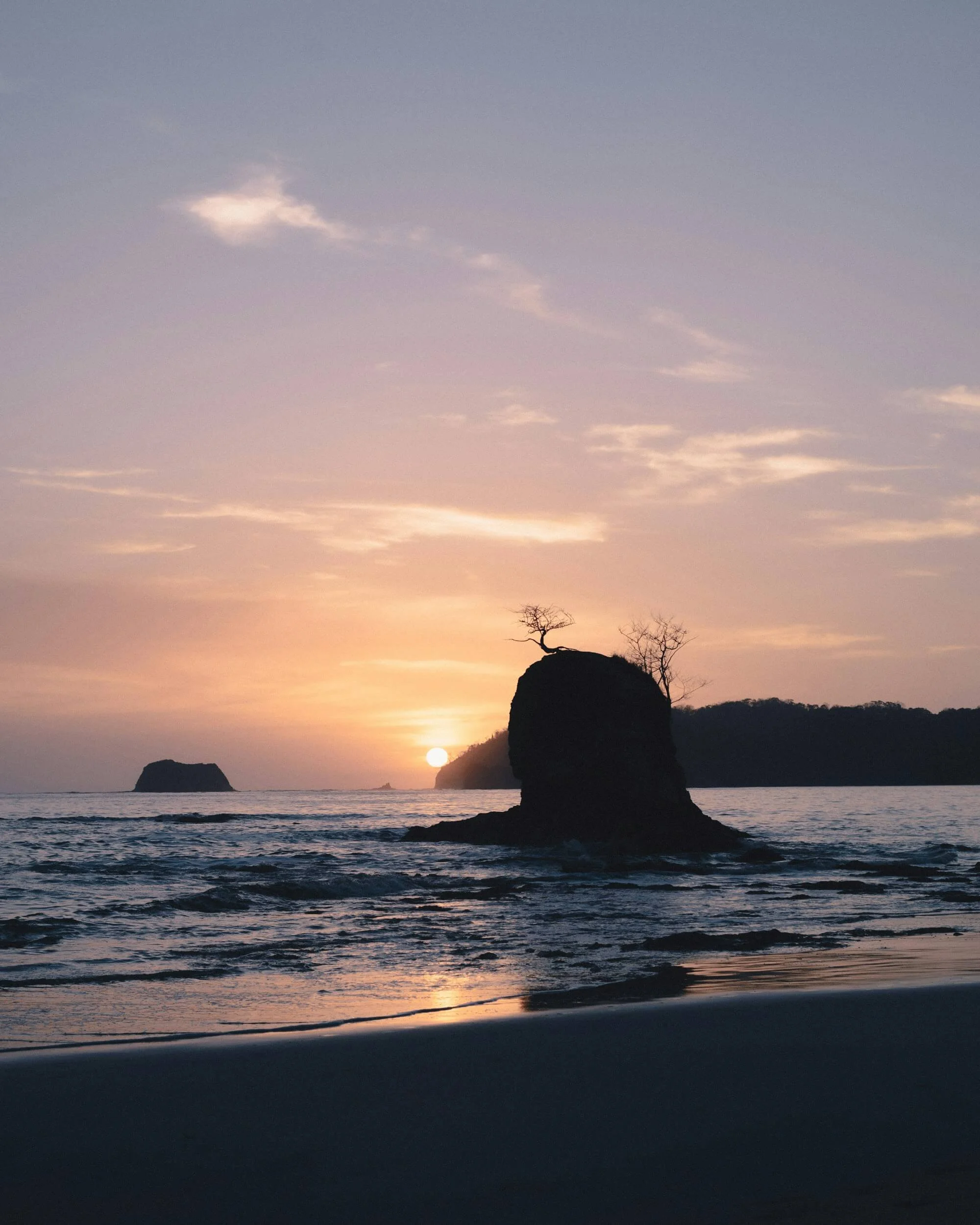 A sunset over the ocean with a large rock formation and two leafless trees on top, silhouetted against the colorful sky.