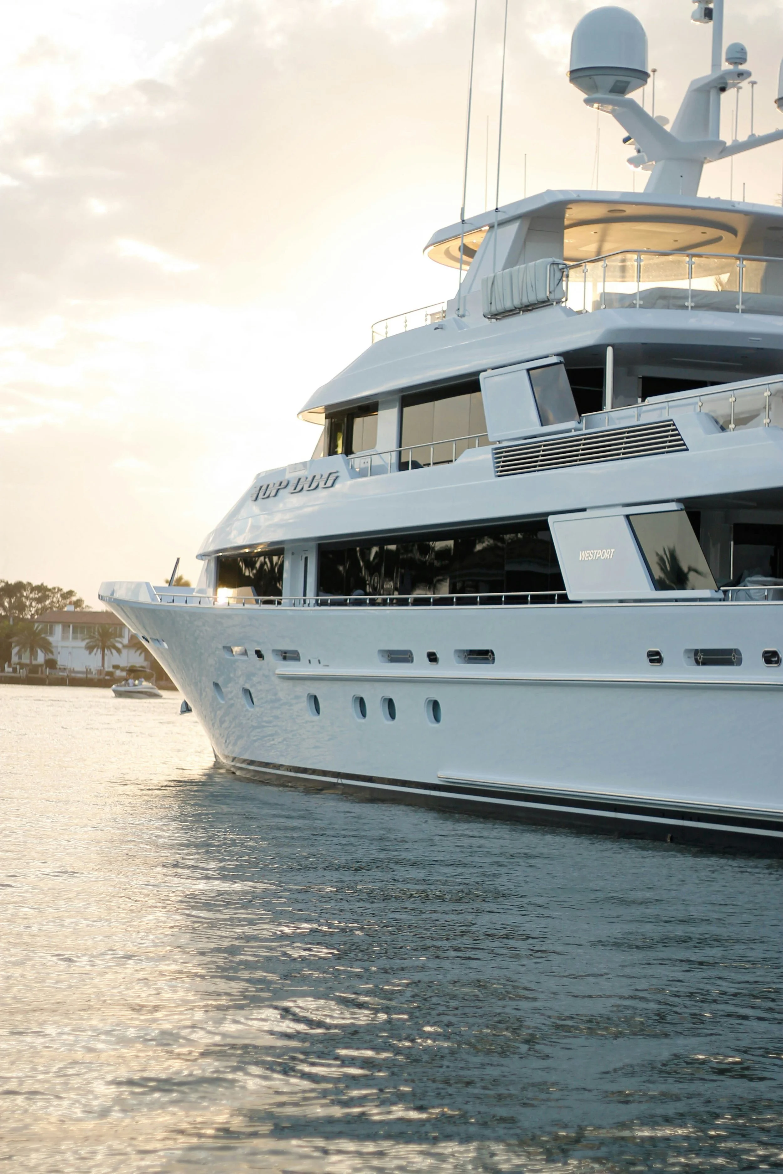 A luxury yacht docked at a marina during sunset with water reflecting the sky and some trees in the background.