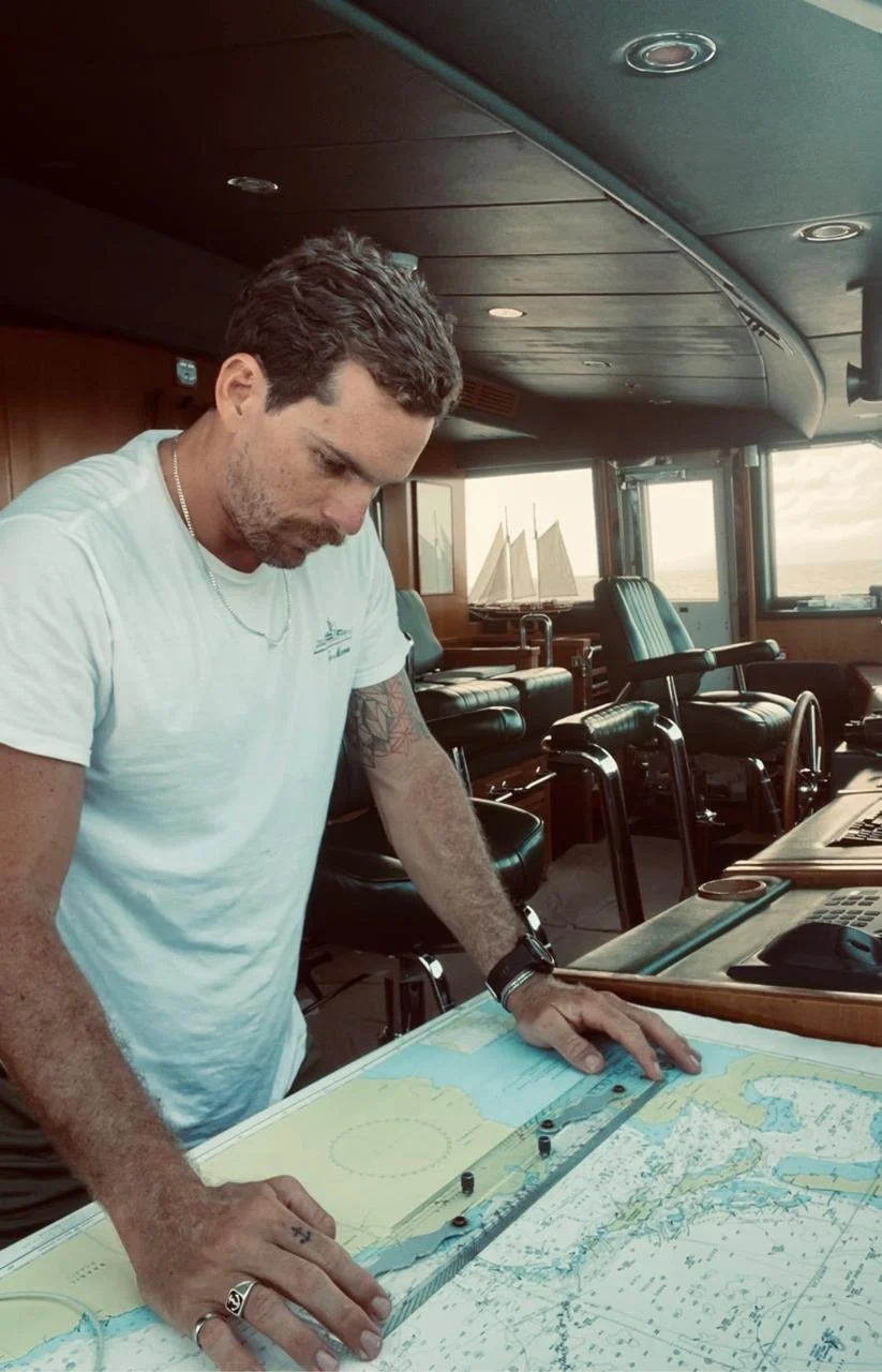 Man studying nautical charts on a ship's bridge with model sailboats on the window ledge and ship controls around him.