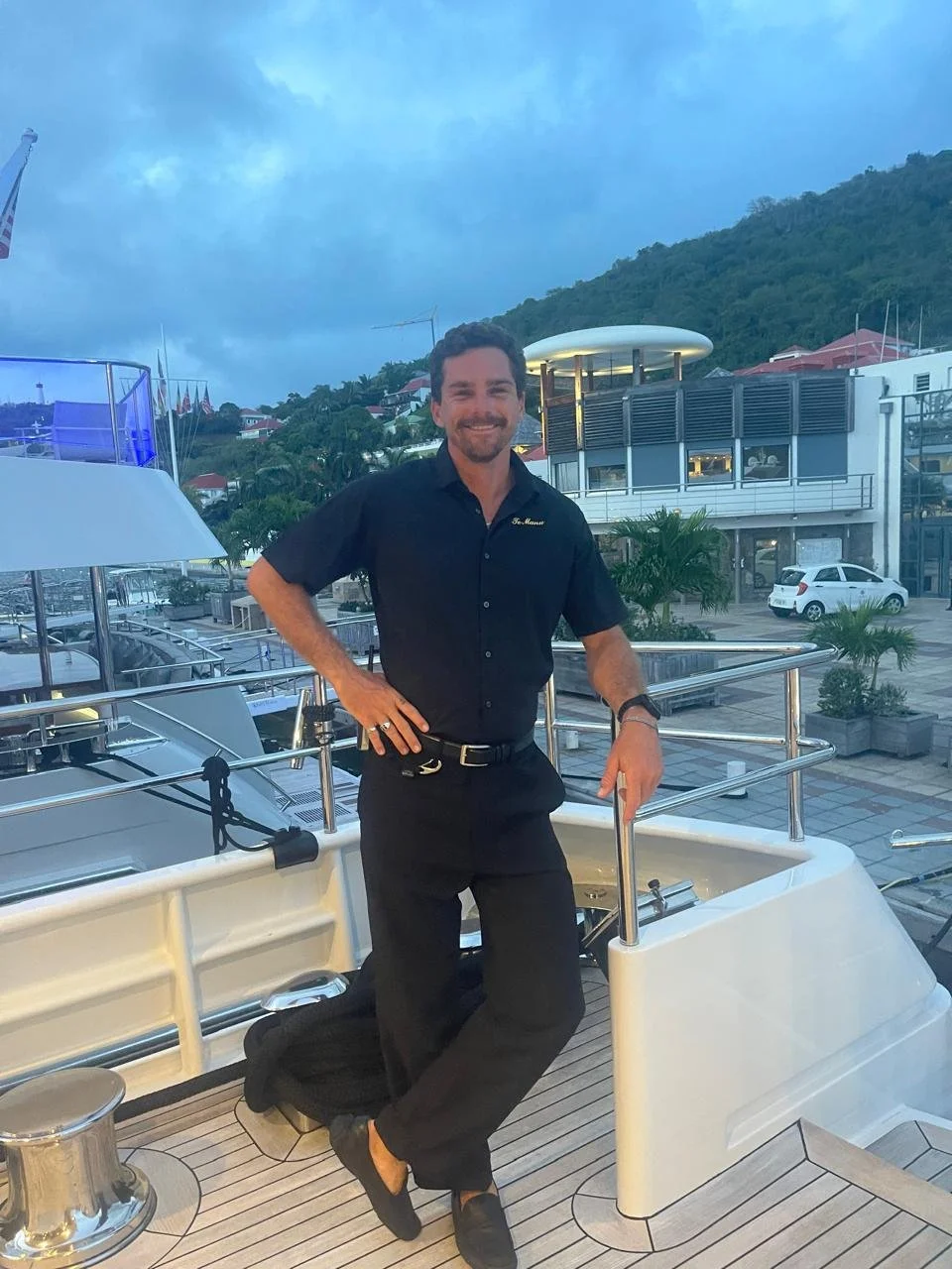 A man in a black uniform kneeling on one knee on a yacht deck, smiling, with a marina and hillside in the background during evening.