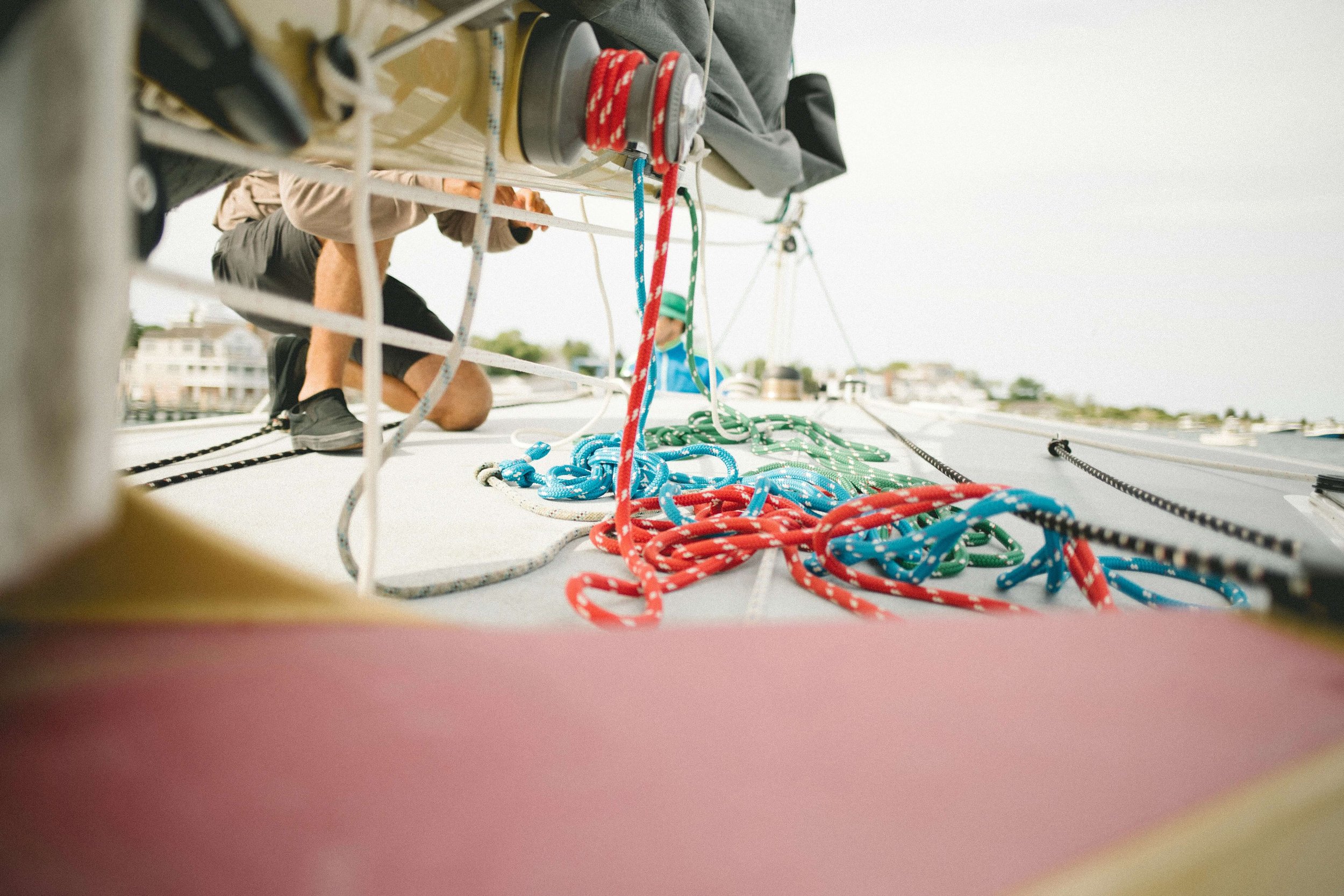 A person working on a boat's rigging, surrounded by colorful ropes, with an overcast sky and waterfront houses in the background.