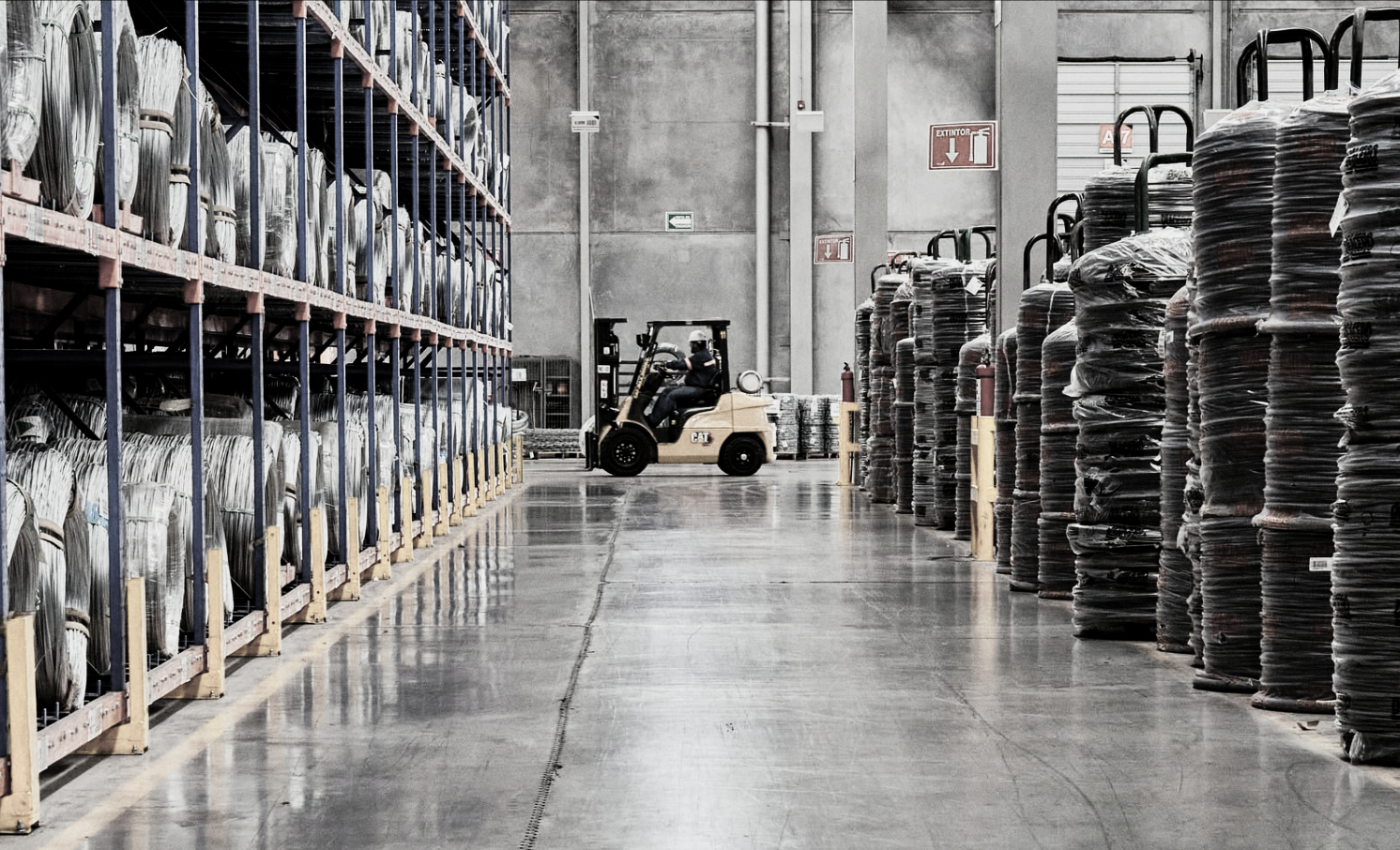 Warehouse aisle with metal shelves stocked with large bags on the left and pallets of rolled materials on the right. A forklift is moving in the background.