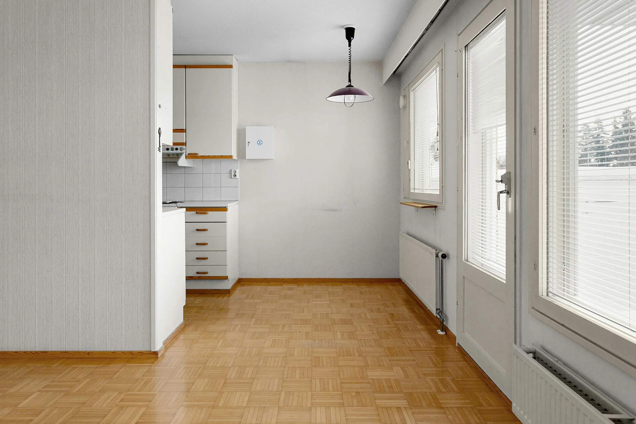 Empty room with hardwood flooring, white walls, and multiple windows with blinds. There is a ceiling light fixture hanging from the ceiling, and a radiator is installed beneath the windows.