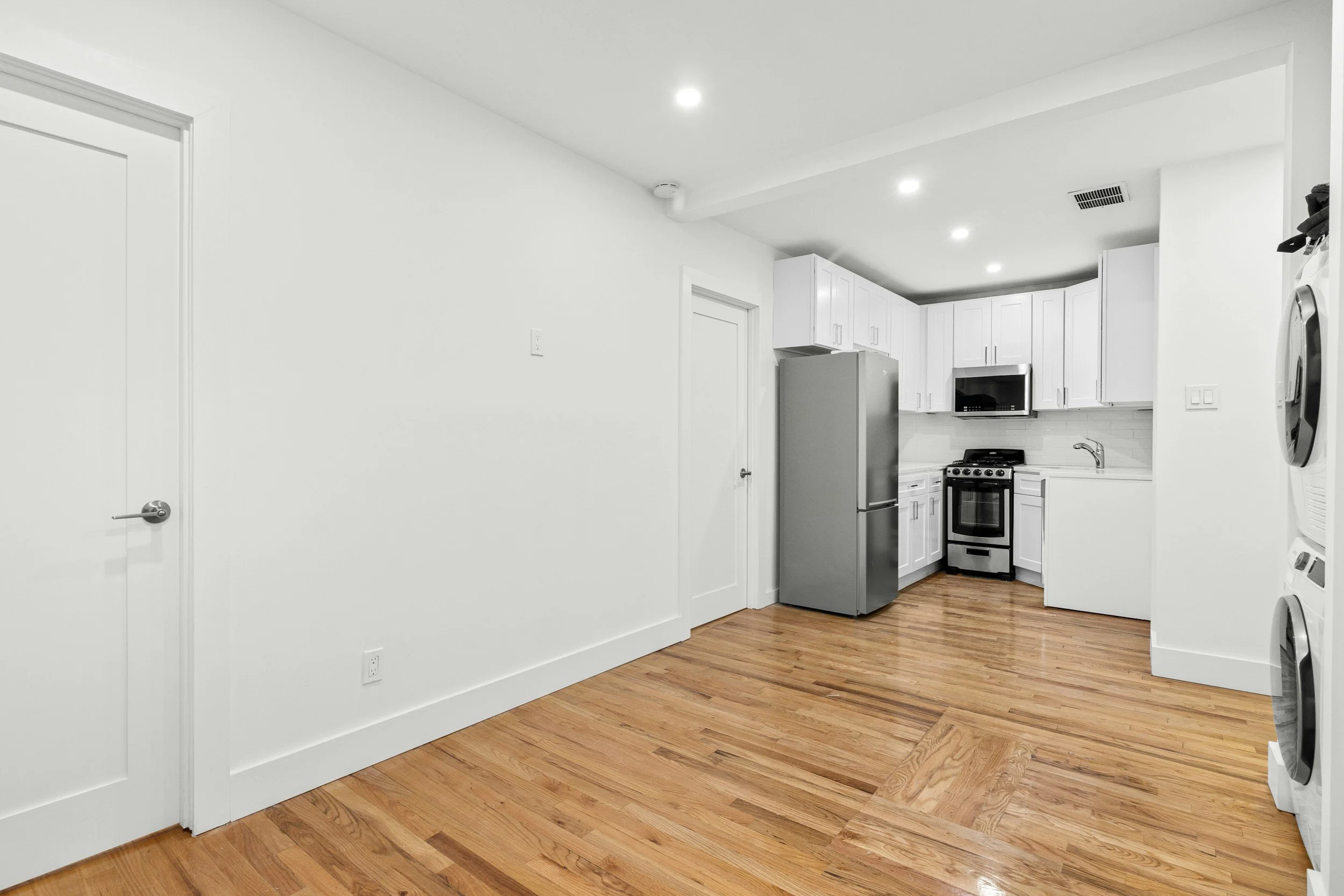 Interior view of a modern kitchen with white cabinets, stainless steel refrigerator and oven, microwave, white walls, hardwood floors, and recessed ceiling lights.