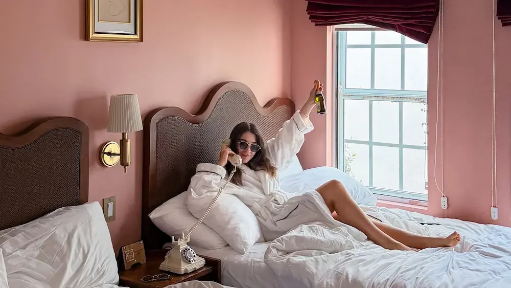 Woman in a white bathrobe sitting on a bed, talking on a landline phone with sunglasses on, holding a beer in one hand, in a pink-walled hotel room with a window and dark red curtains.