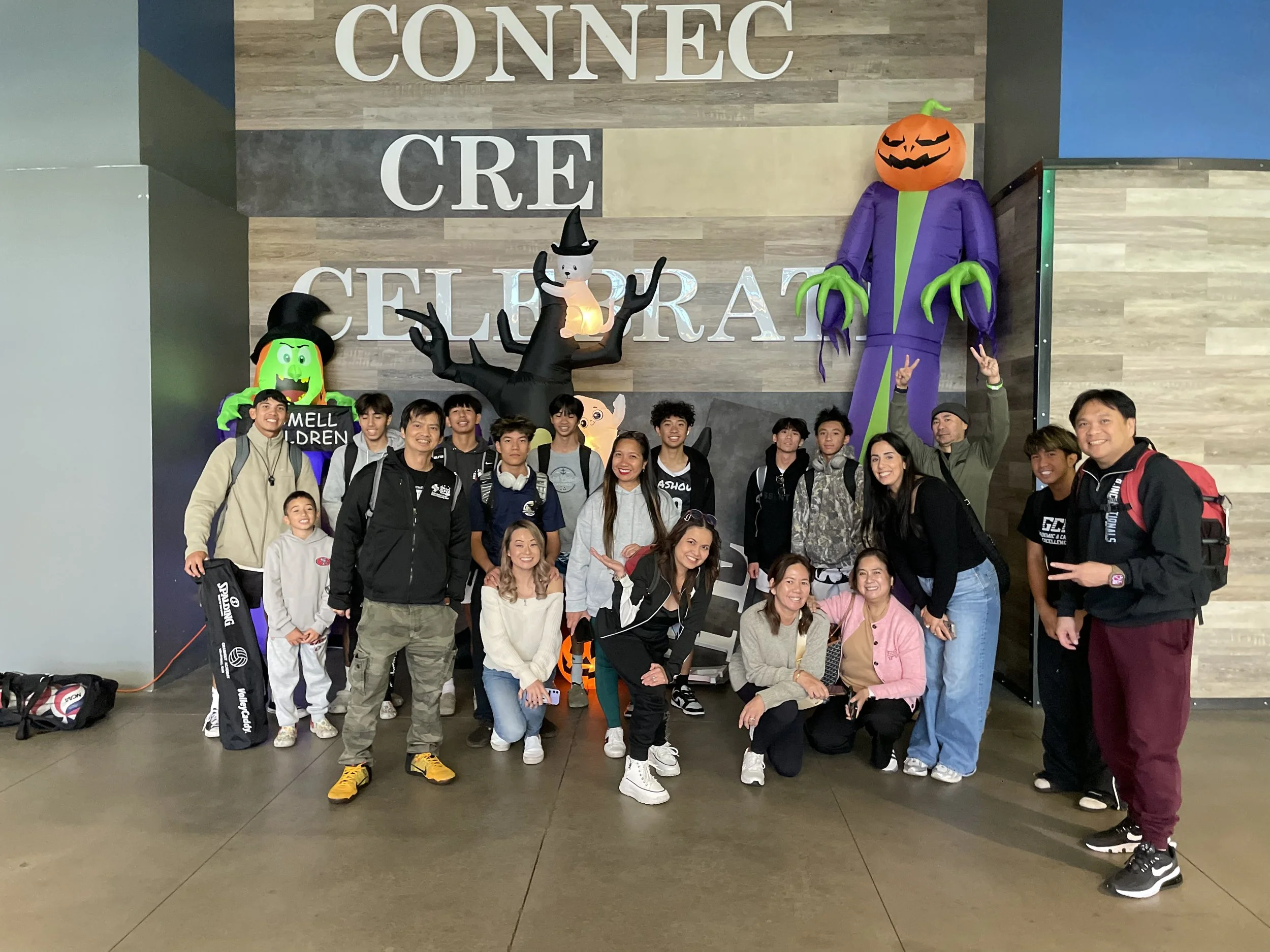 A group of people posing in front of a wall with large Halloween decorations, including a pumpkin-headed figure and a black cat. The background features a wooden wall with the words 'Connect Create Celebrate' in large white letters. Everyone appears 