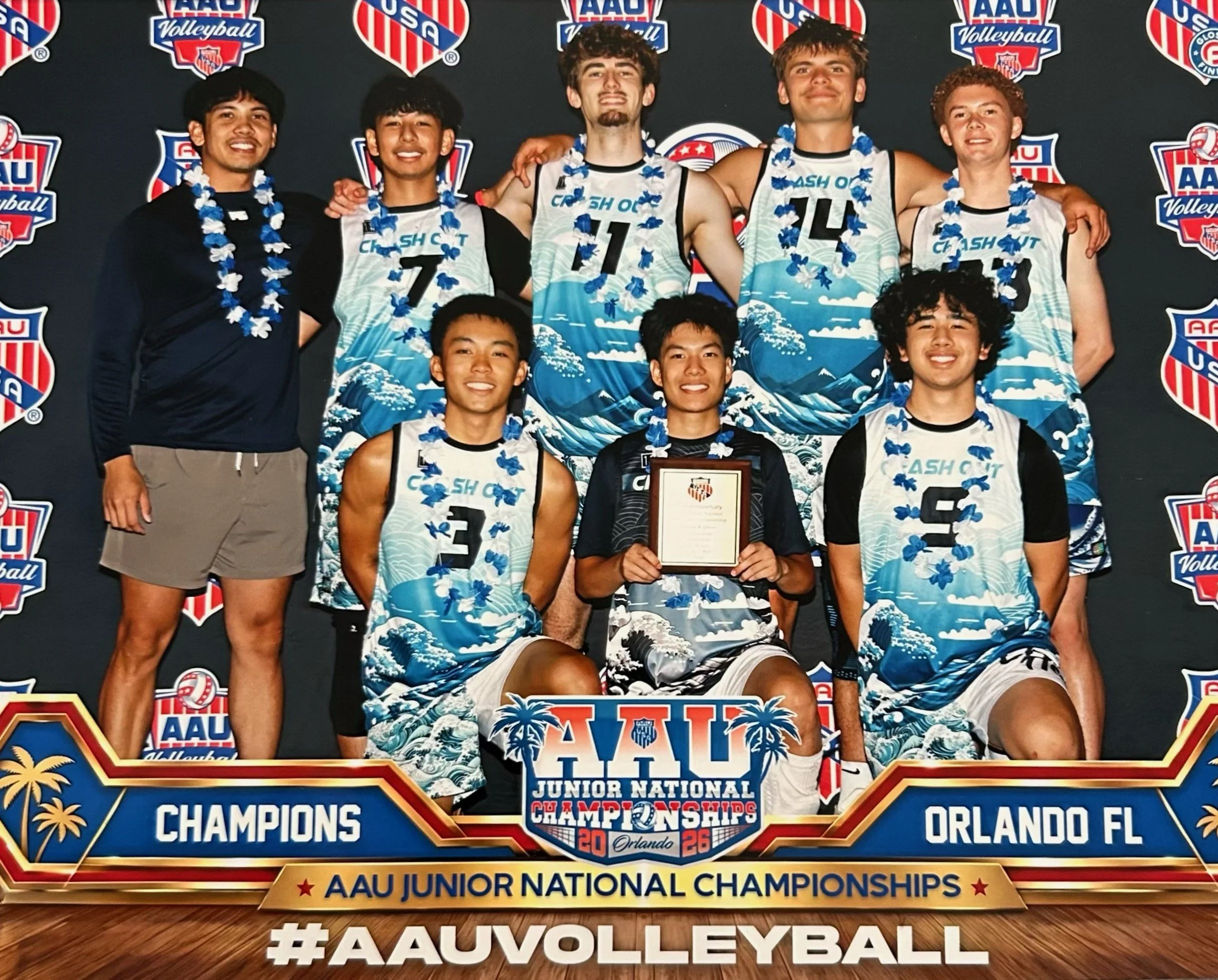 Young male volleyball players and coach celebrating after winning the AAU Junior National Volleyball Championships in Orlando, Florida, in 2026. The team members are wearing matching blue and white uniforms with a wave design and leis, standing in fr