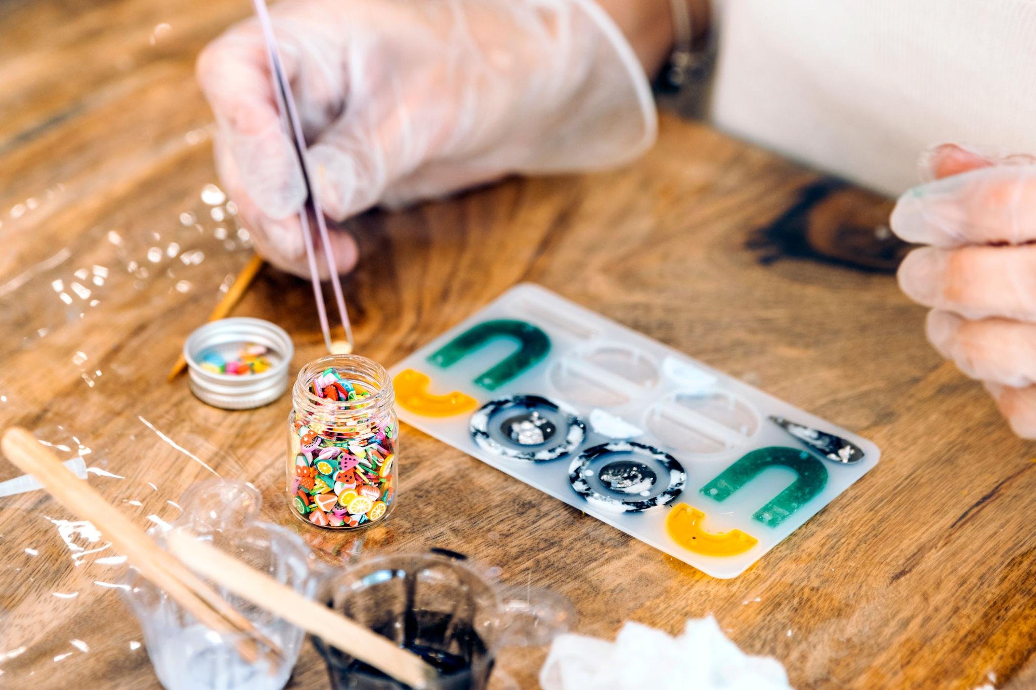 Close-up of a sand art craft project being made with small colorful seed beads in a plastic container, wooden sticks, and multi-color paints on a wooden table.