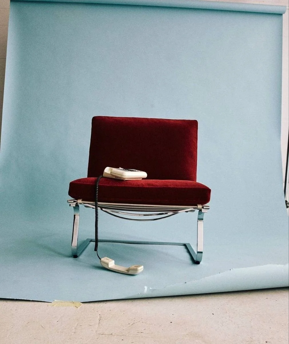A red velvet chair with a beige rotary telephone and cord resting on the seat, set against a light blue backdrop.
