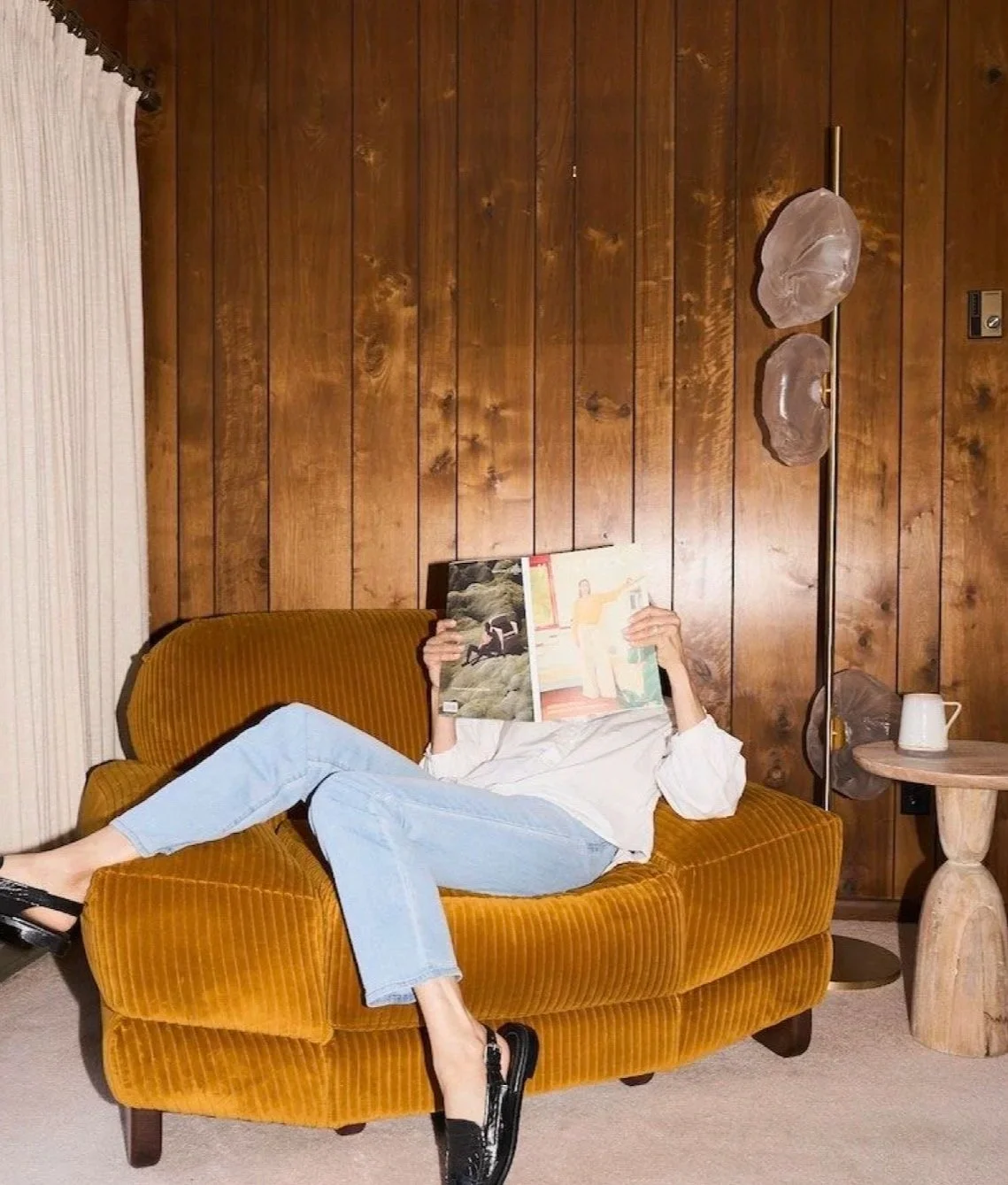 Person lying on a mustard yellow couch, holding an open magazine to hide their face, in a room with wood-paneled walls, a floor lamp with decorative glass shades, and a small round wooden side table with a white pitcher.