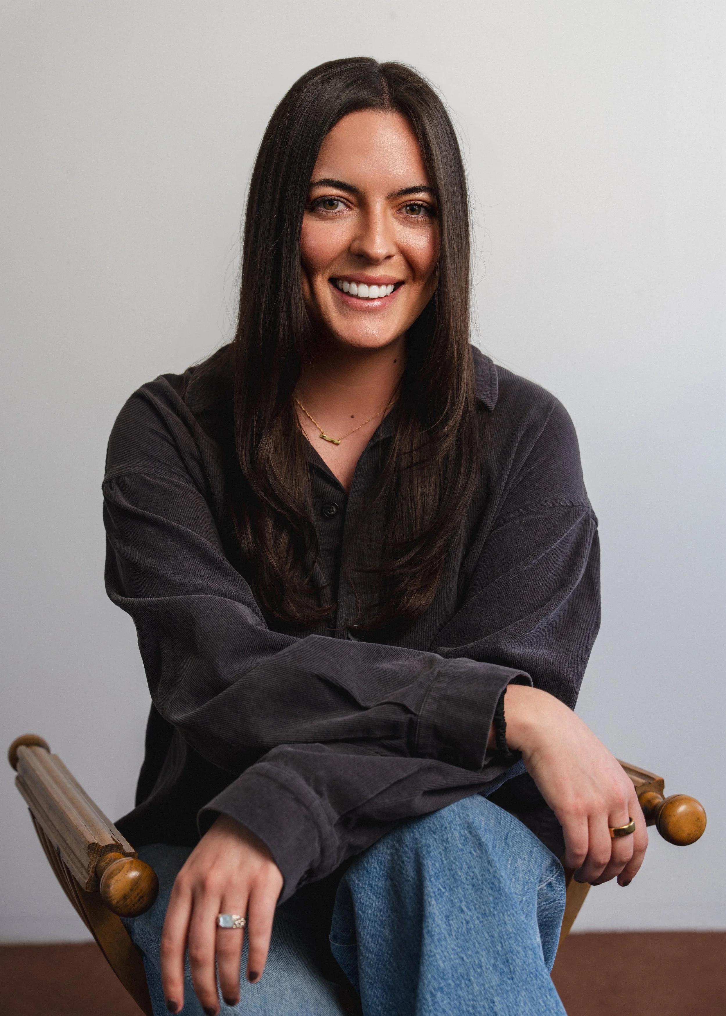 A woman with long dark brown hair smiling, sitting on a wooden chair with arms crossed, wearing a dark button-up shirt, blue jeans, a gold necklace, and rings, against a plain white background.
