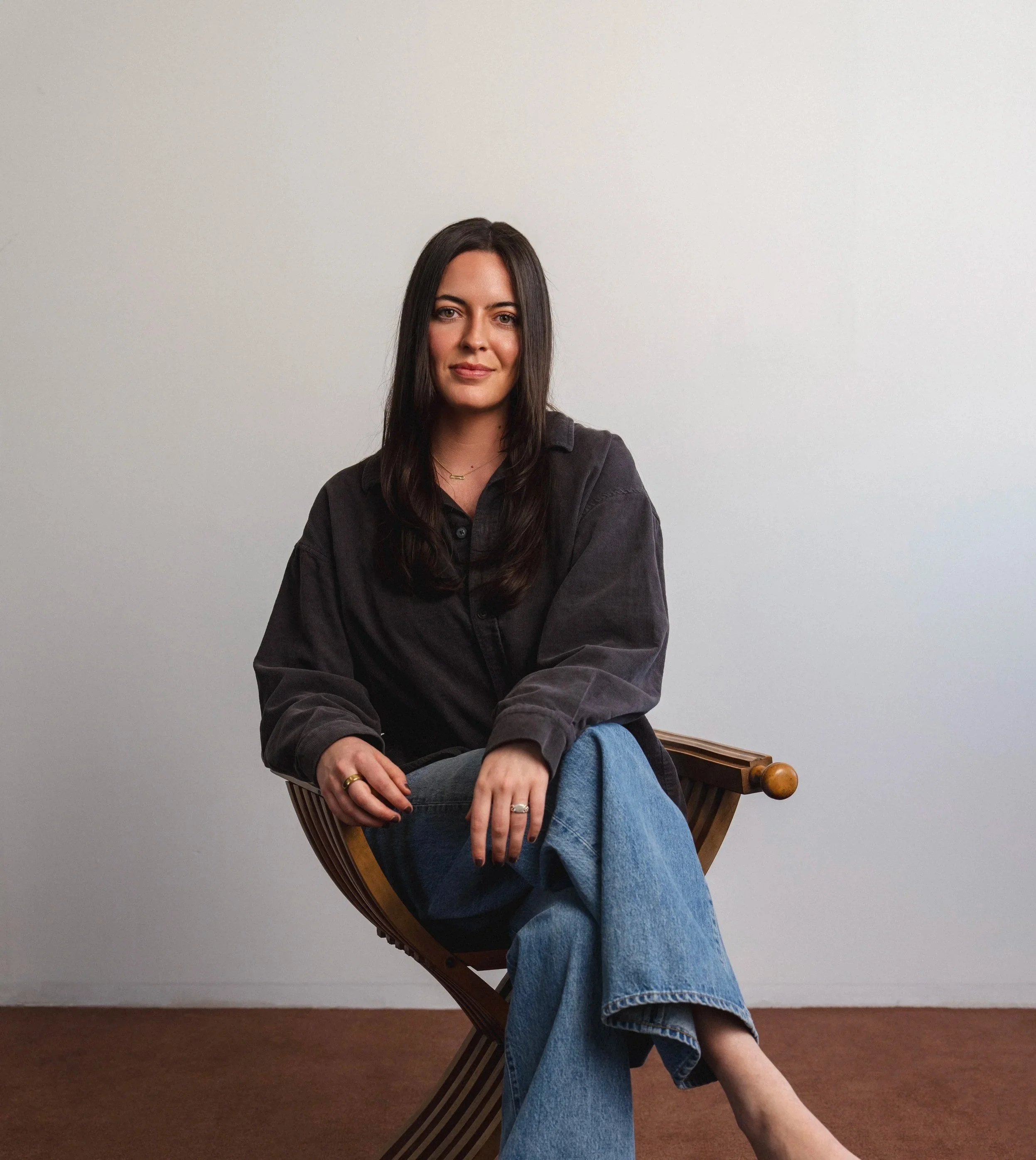 A woman with long dark hair is sitting on a wooden chair positioned sideways in front of a plain white wall, wearing a black shirt, light blue jeans, and several rings, with a relaxed expression.