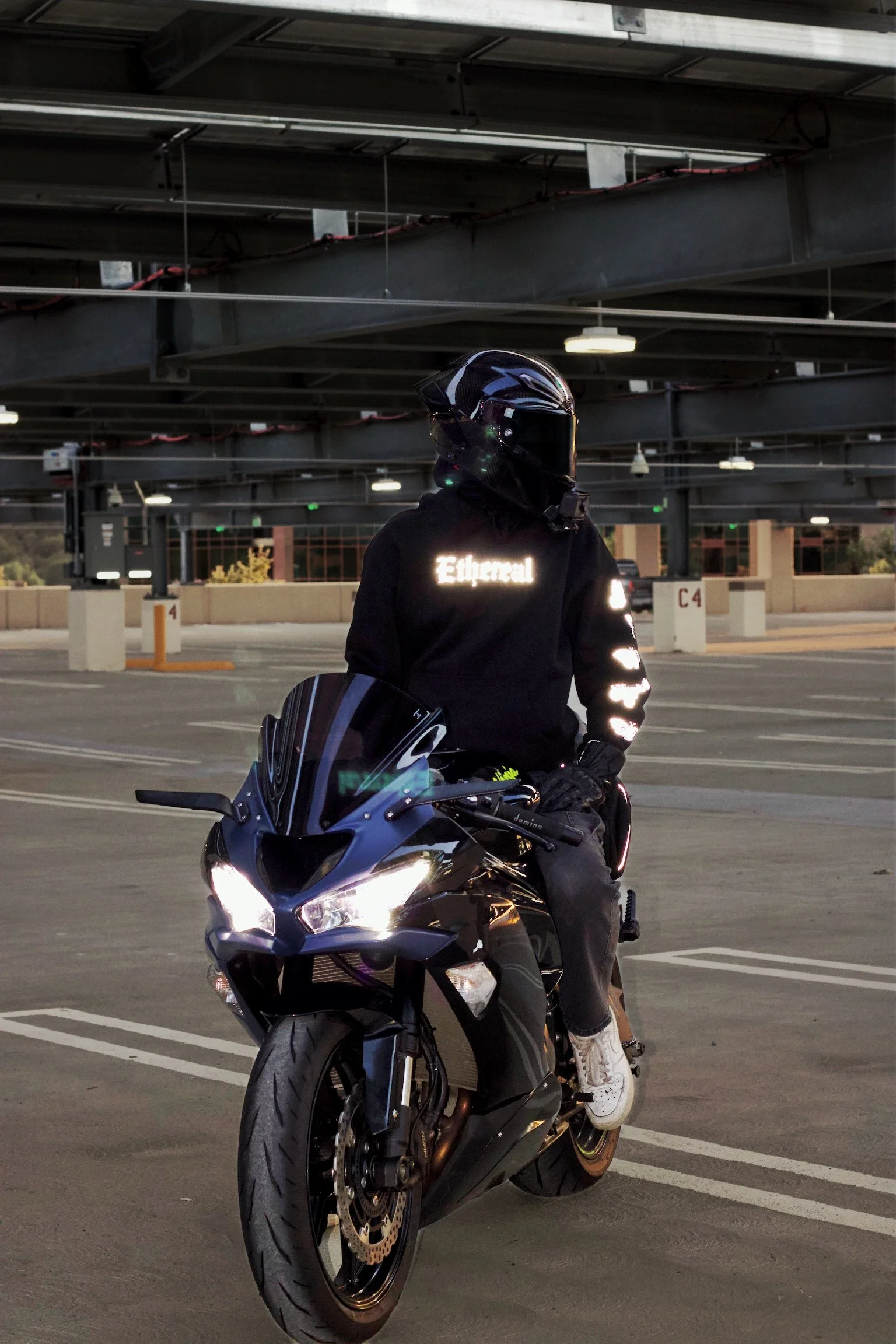Person wearing motorcycle gear, including a reflective jacket with the word 'Eternal', sitting on a black motorcycle in a parking garage, with a helmet on and bright headlights.
