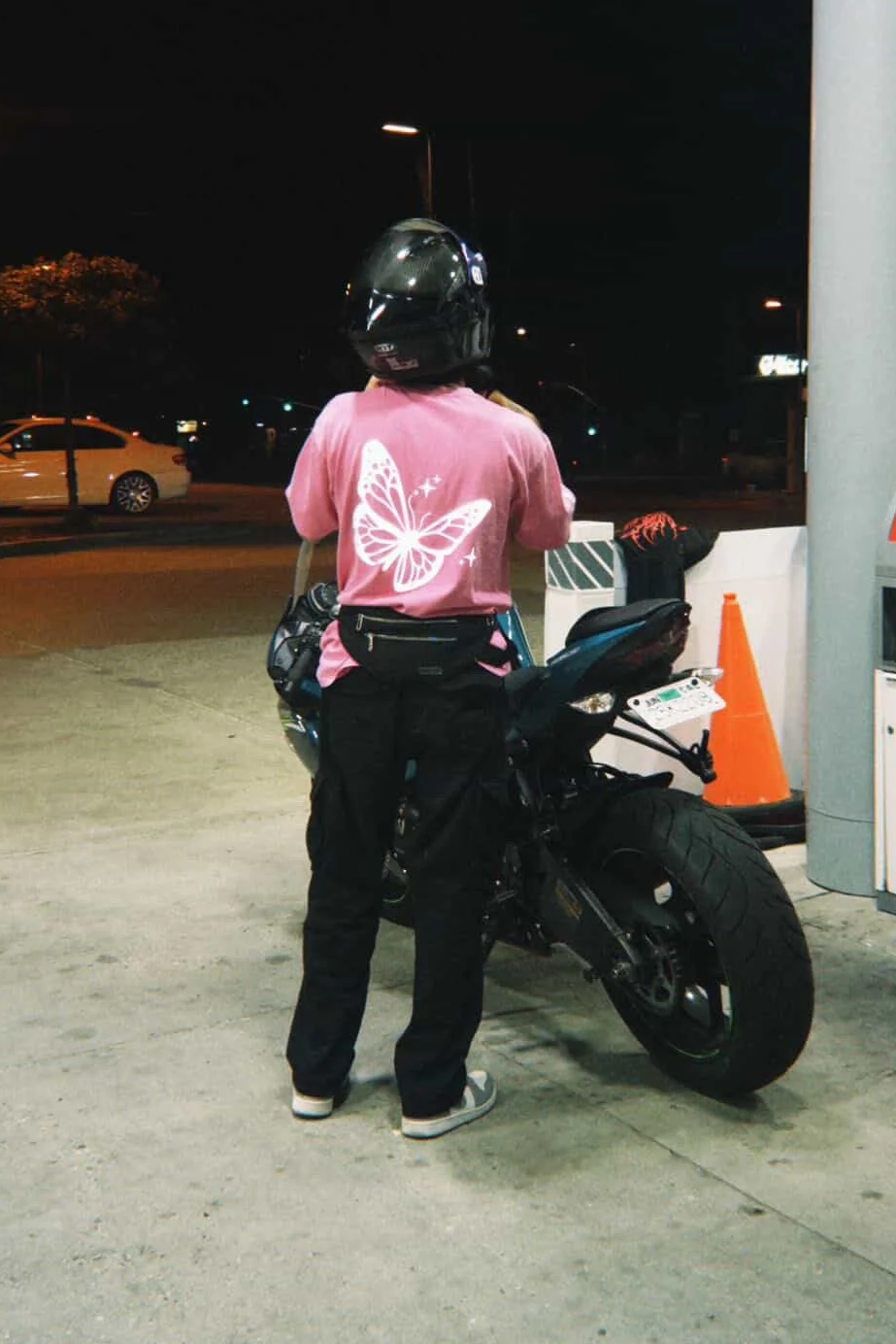 Person standing next to a black motorcycle at night, wearing a pink shirt with a butterfly design, black pants, gray shoes, and a black helmet.