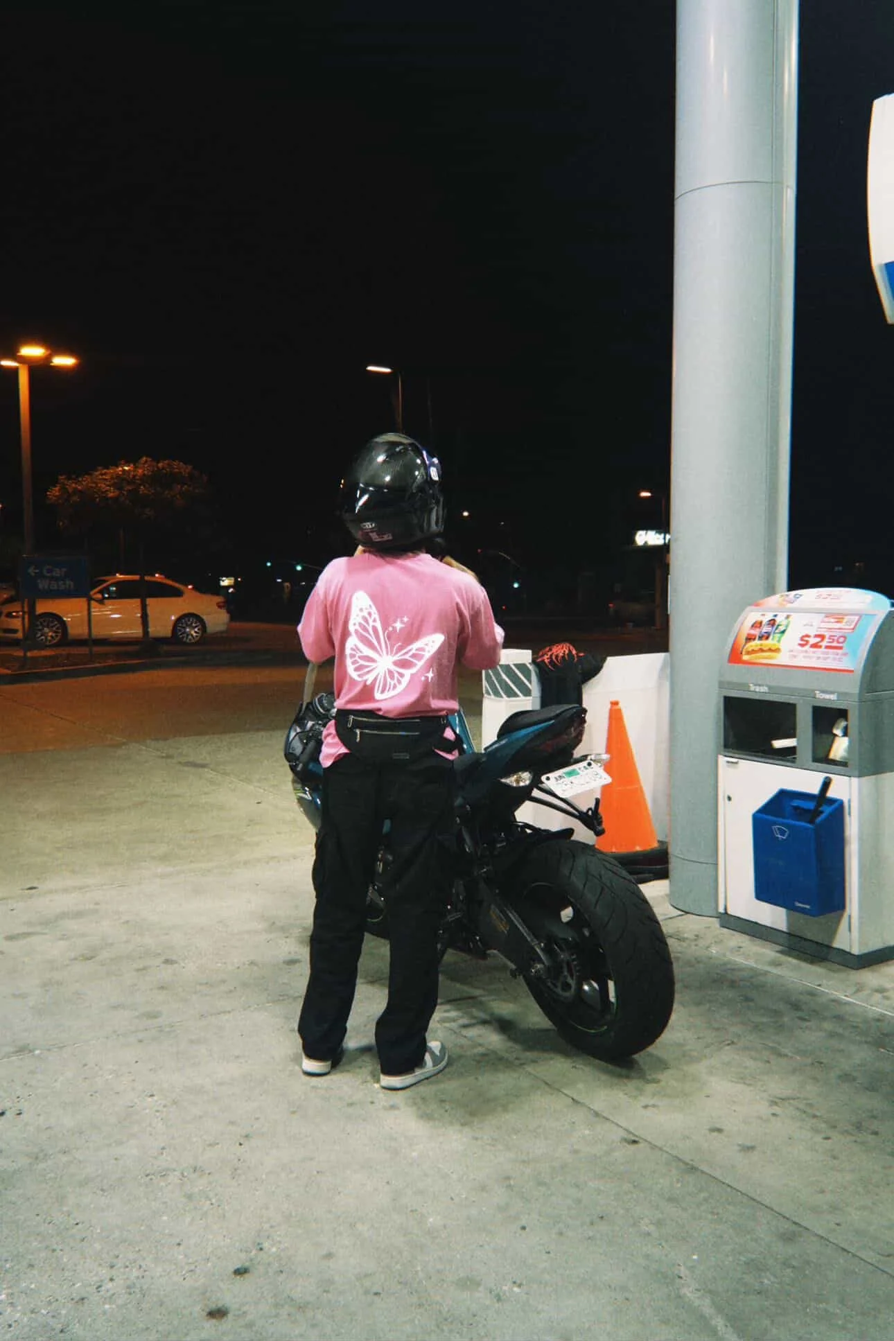 Person in pink shirt with butterfly graphic and black helmet standing next to black motorcycle at gas station at night.