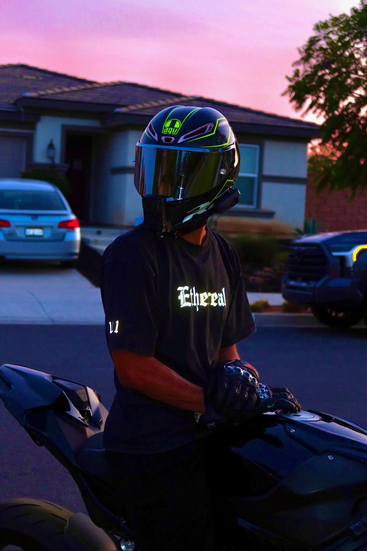 A person wearing a black AGV motorcycle helmet with a reflective visor, a black T-shirt with white text, and gloves, sitting on a black motorcycle during sunset in a suburban neighborhood with parked cars and a house in the background.