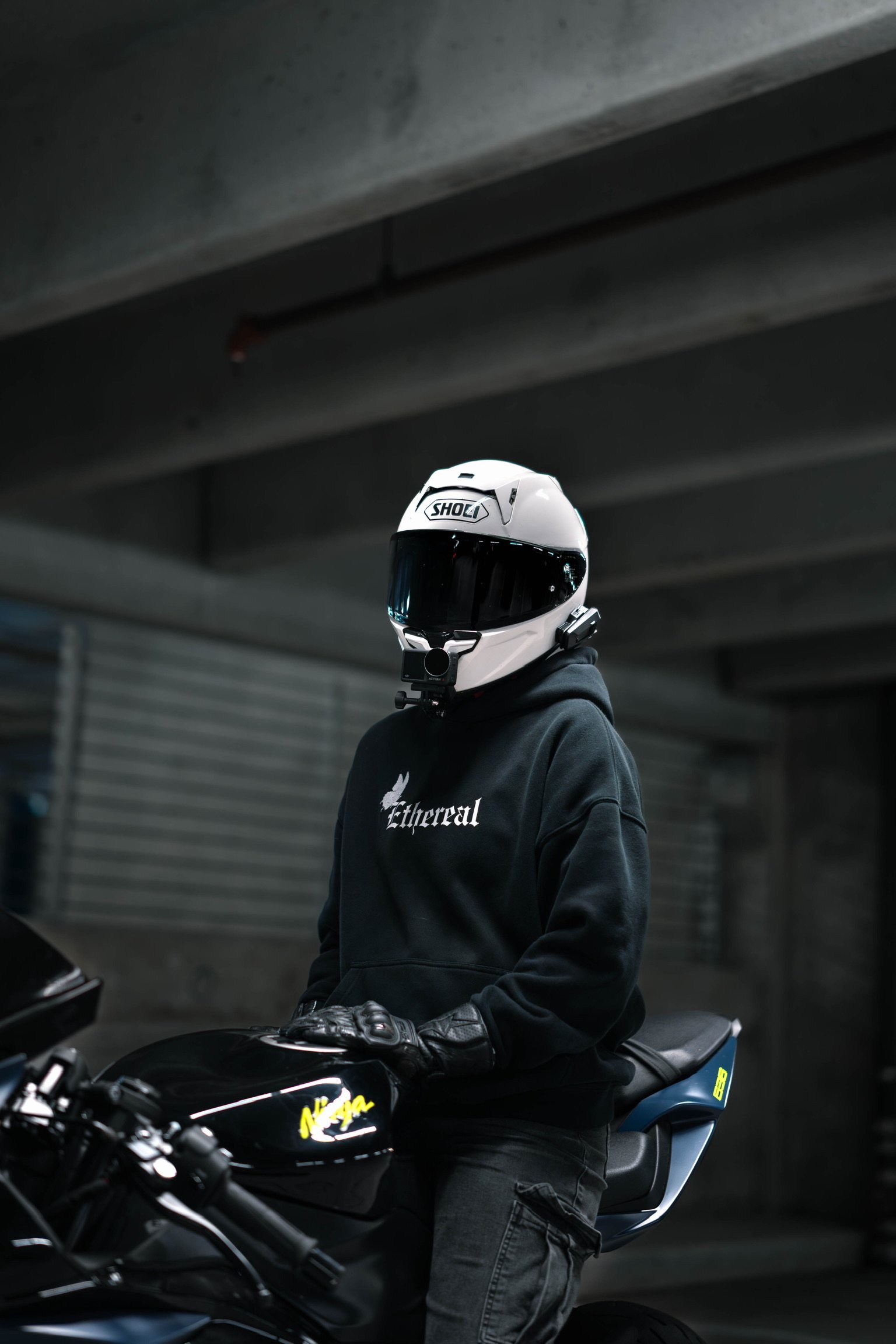 A person wearing a Shoei helmet and black gloves sitting on a black motorcycle in a dark garage, with a large closed metal rolling door in the background.