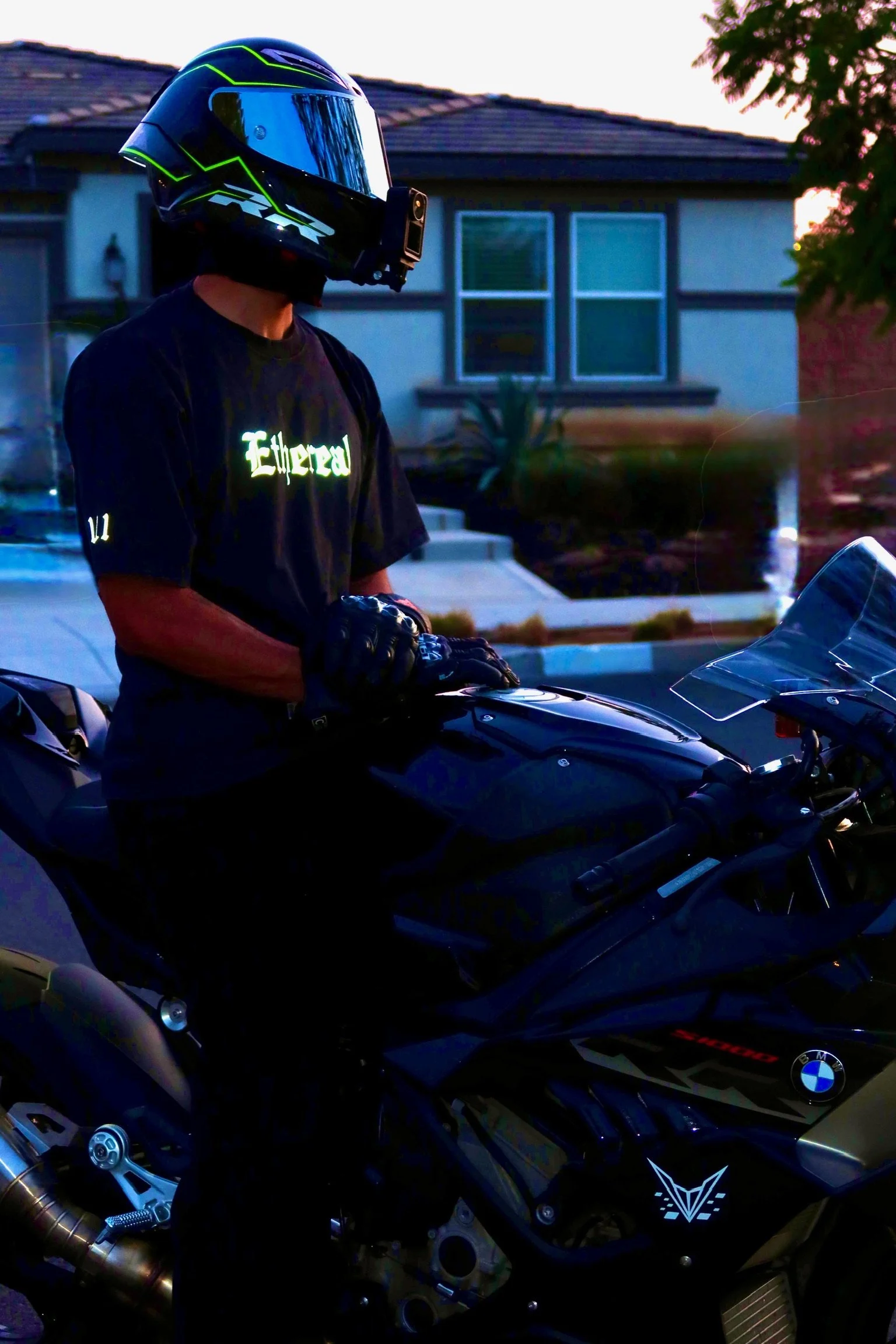A person wearing a black helmet with a blue visor and a GoPro camera attached, standing next to a black BMW motorcycle with BMW logos, in a residential neighborhood during sunset.