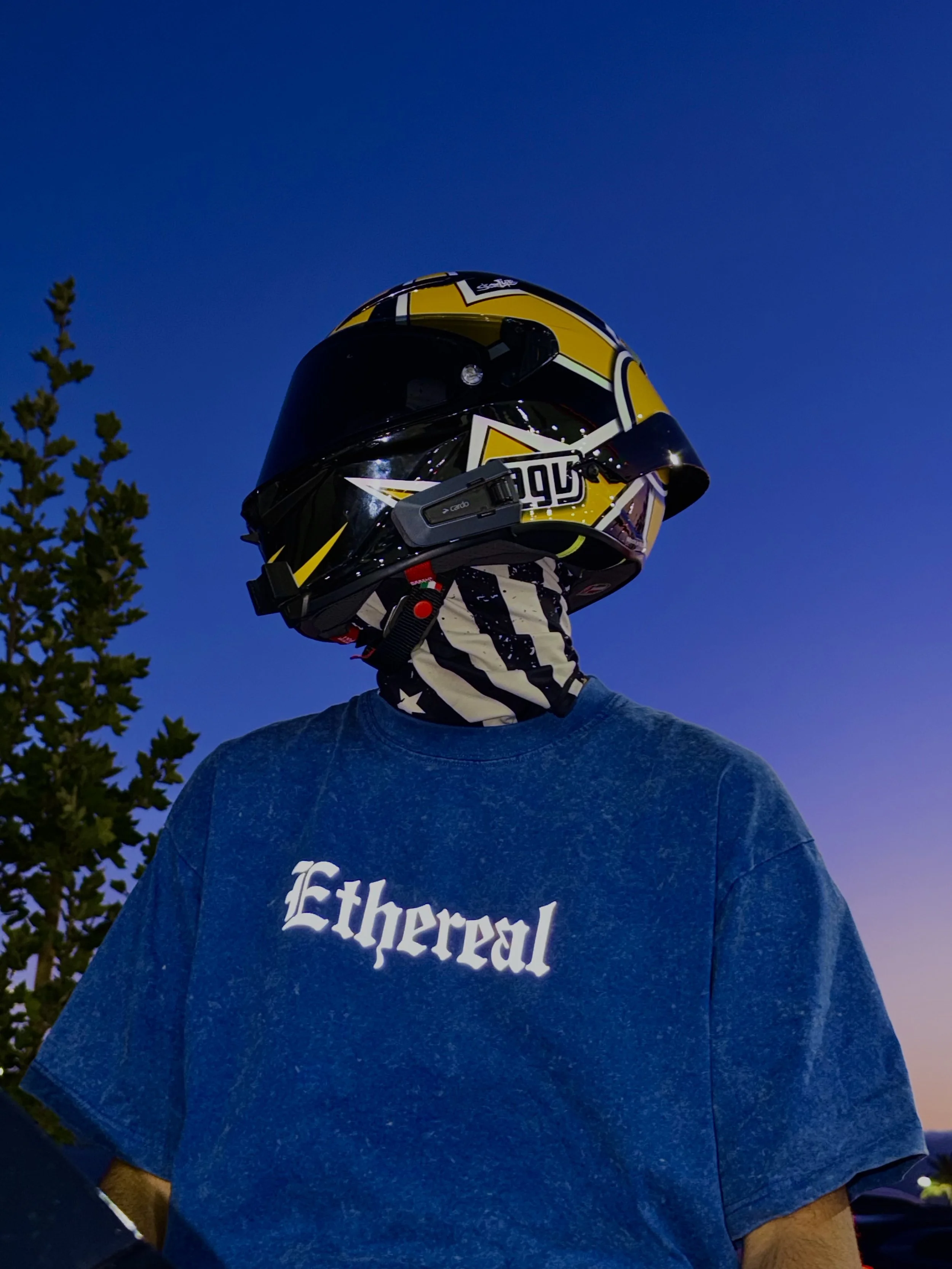 A person wearing a black and yellow motorcycle helmet, a blue T-shirt with 'Ethereal' written on it, and a black and white bandana with stars. The person is standing outdoors during twilight with a tree and a colorful sky in the background.